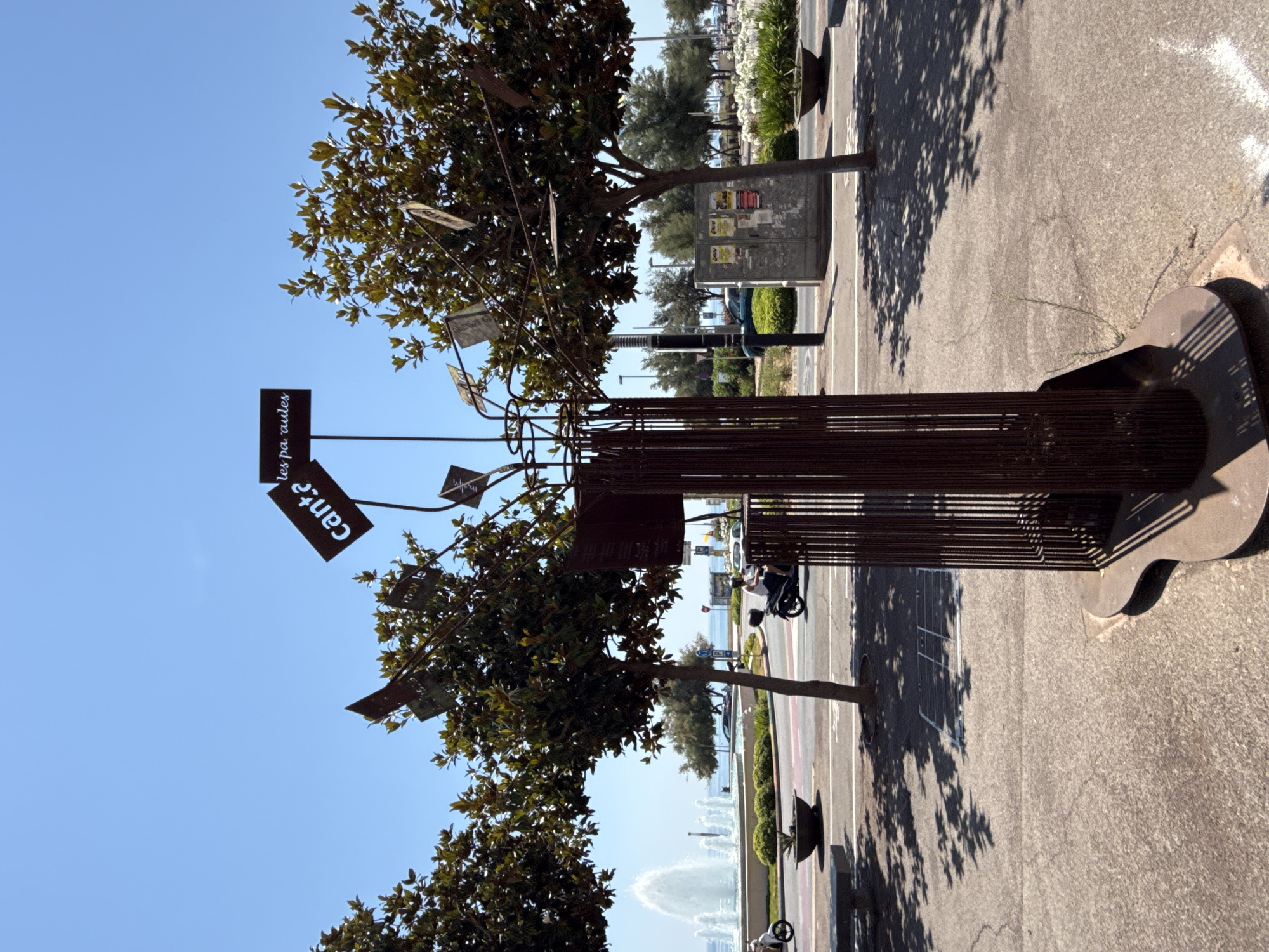 The image depicts an outdoor sculpture made of dark metal, resembling a tree with multiple branches. Attached to the branches are several rectangular metal plates, some of which have text on them. One plate reads "canta" in white, bold letters. Another plate, further to the right and higher up, has the text "les pa ules" written in a smaller font.
The sculpture stands on a paved area, casting shadows from the surrounding trees. In the background, a street with traffic, including a scooter with a rider, is visible. Beyond the street, there's a large circular fountain with water jets. Further in the distance, the sea is visible, with a clear blue sky overhead. Two trees with lush green foliage frame the central sculpture. To the right of the sculpture, a light-colored electrical box is mounted with several posters attached to it. The overall environment suggests a public urban space, likely a promenade or park area near the coast. The presence of the sea, fountain, and street activity indicates a location in a coastal town. This scene is located in the city of Blanes, Spain.