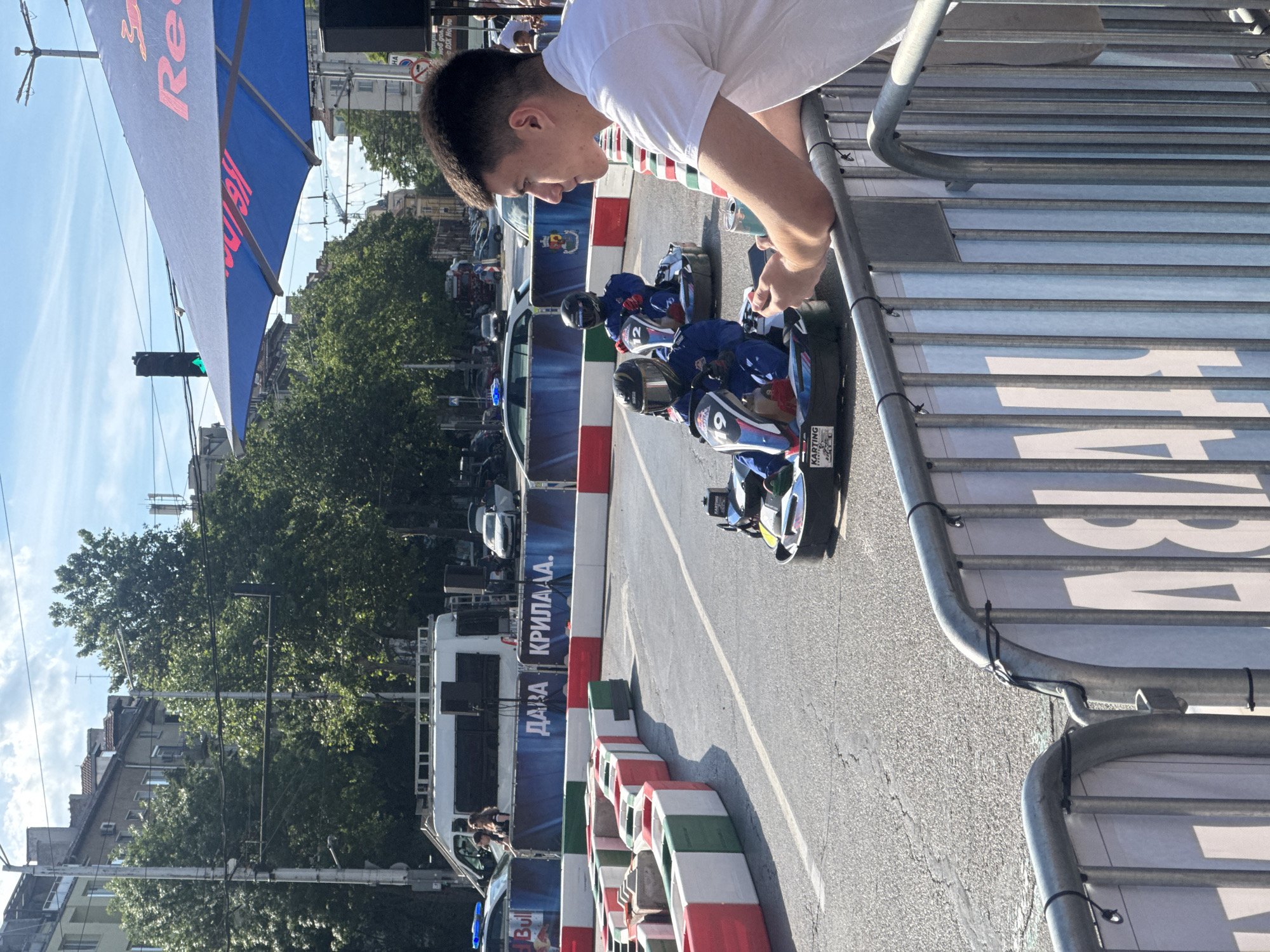 A young man in a white t-shirt is looking over a metal fence at a go-kart race. The go-karts are blue and white with red accents, and the drivers are wearing helmets and racing suits. The race track is lined with red and white barriers. In the background, there are trees, buildings, and a Red Bull umbrella. A sign in Cyrillic reading "ДАВА КРИЛА А.А." is visible. The location is in Sofia, Bulgaria.