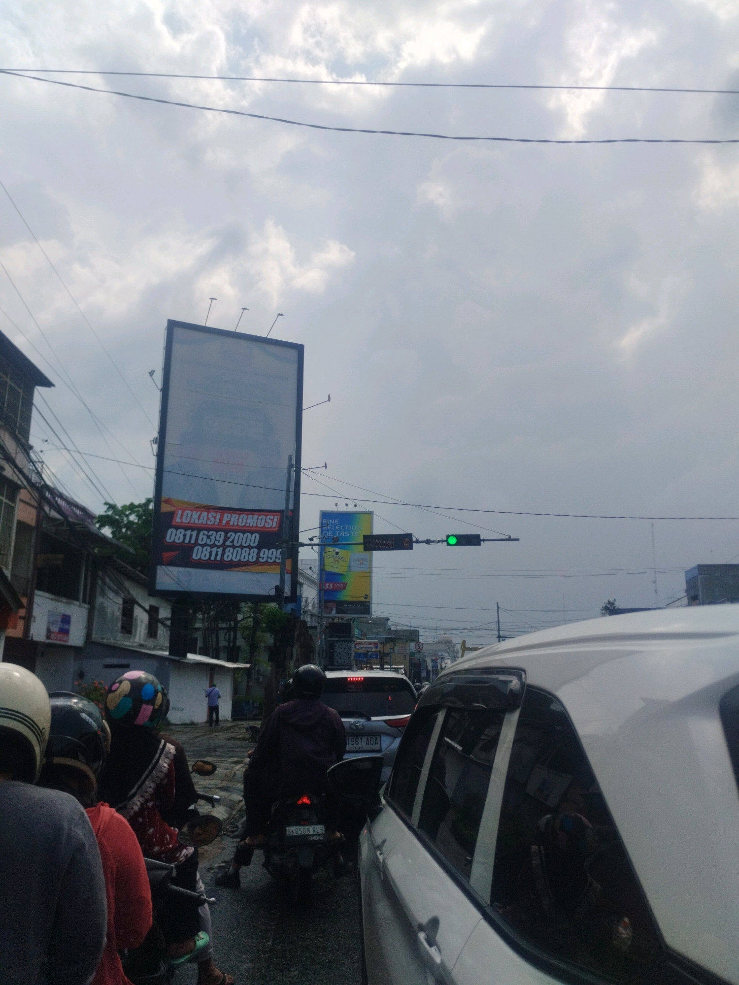In the City of Medan, Indonesia, under a cloudy sky, a traffic jam is depicted. Several motorcycles and a white car are stopped on a wet street. On the left side of the image, a billboard displays advertisements with text including "LOKASI PROMOSI" and phone numbers. Further down the street, a traffic light shows a green signal. People on motorcycles are wearing helmets of various colors and designs. One person on a motorcycle is wearing a purple headscarf and a red and white patterned outfit, while another is wearing a dark hooded jacket. The white car in the foreground partially obstructs the view of the road ahead.