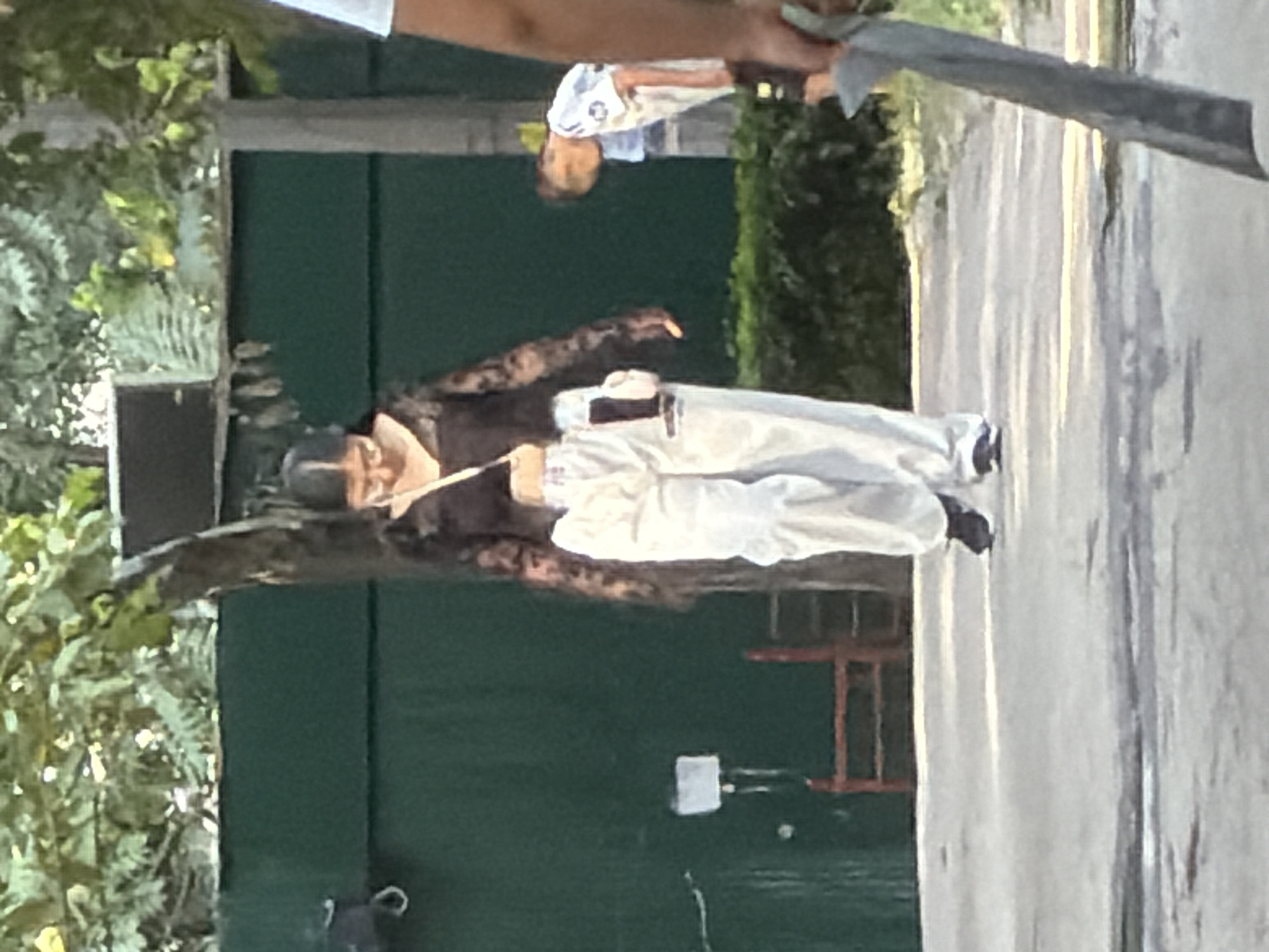 An outdoor scene shows a woman in the foreground, wearing a black sheer long-sleeved top, white pants, and black shoes. She has a white purse with a pink detail slung across her body. She is looking down and walking. Behind her, to the right, a person in a white and blue shirt is bending over. The background features a dark green wall and trees. The ground is a light-colored path. In the lower right corner, a person's arm is visible, holding a large, flat, gray object.