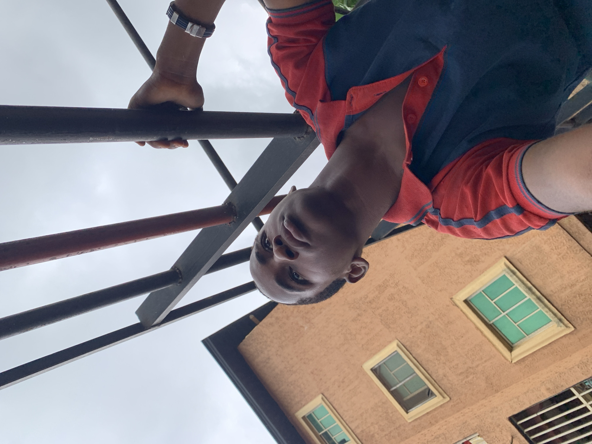 A young Black male is pictured from a low angle, looking upwards. He is wearing a red and navy blue polo shirt and a bracelet on his left wrist. His left hand is holding onto a metal bar, which is part of a structure that appears to be outdoors, possibly a playground or scaffolding. Behind him, to the left, is a peach-colored building with several windows, some of which have green panes. The sky is overcast. The scene is set in Nkpor, Nigeria.