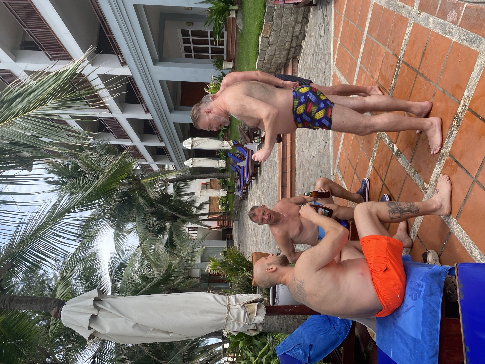 In Mũi Né, Vietnam, three men are socializing outdoors near a hotel. The men are wearing swimwear and holding bottles of beer. One man, with a shaved head and a tattoo on his back, sits on a blue lounge chair and holds a beer bottle. Another man, with a receding hairline, sits next to him and also holds a beer bottle. The third man, with a beard and a colorful patterned swimsuit, stands and gestures with a beer bottle. The scene takes place on a paved patio with palm trees and a multi-story hotel in the background.