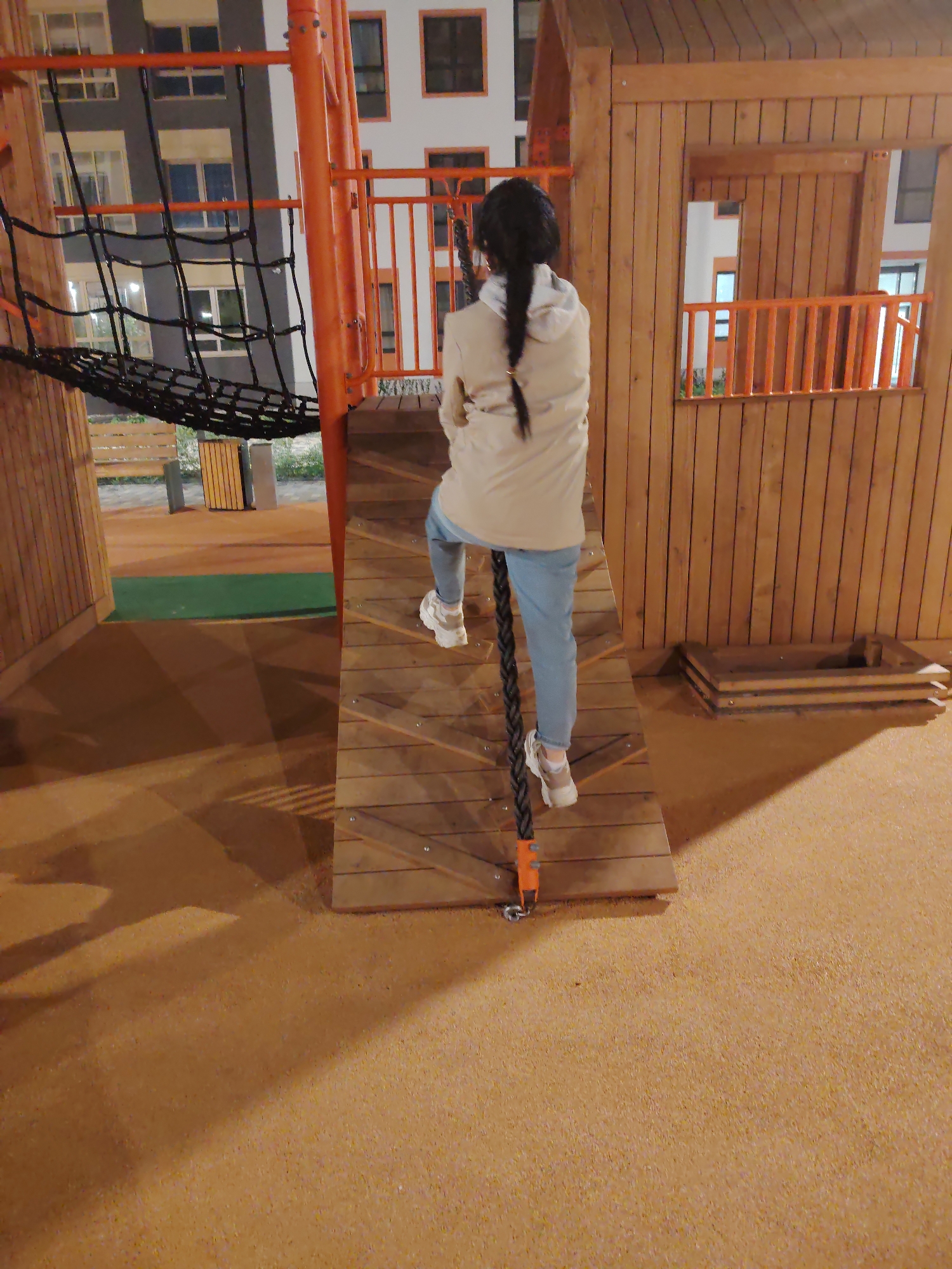 The image shows a young person with dark hair braided down their back, wearing a light beige hoodie and light blue jeans. They are positioned on a wooden climbing ramp with a thick rope, seemingly ascending. The background features a wooden playhouse with a window and orange railings. To the left, an orange metal structure supports a black net. The playground is surfaced with a tan, granular material, and in the background, residential buildings are visible. The scene is lit by artificial lights, suggesting it is nighttime. The overall environment appears to be a modern outdoor playground.