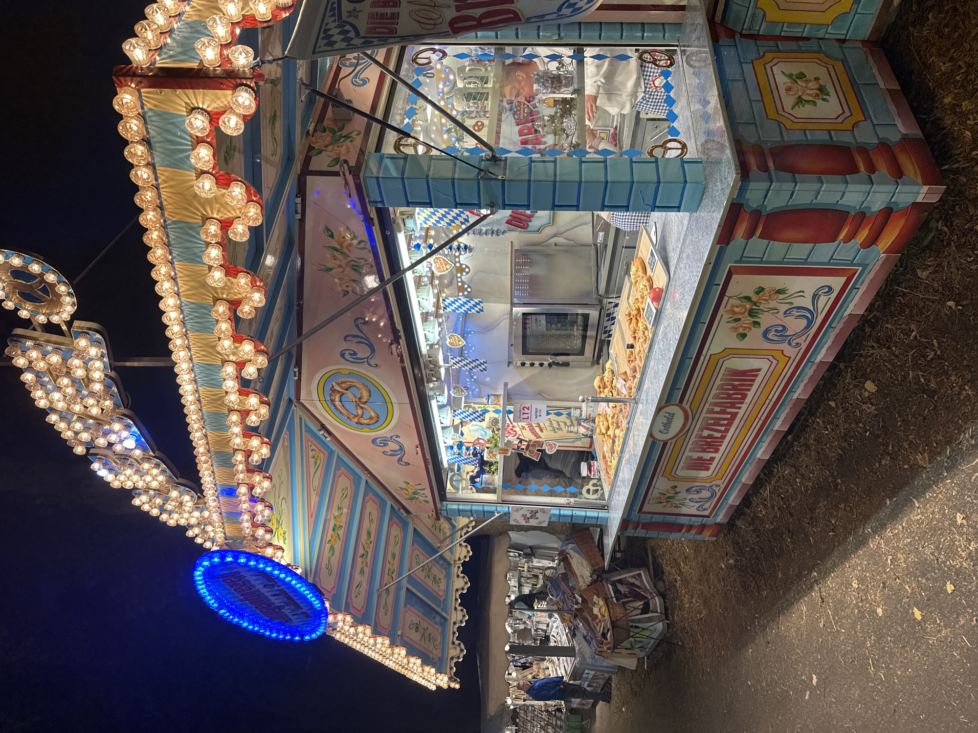 The image captures a festive scene at night, likely at a market or fair in Bruchhausen-Vilsen, Germany, focusing on a pretzel stand. The stand is brightly illuminated by numerous warm-toned lights adorning its elaborate canopy and facade, which is decorated with floral motifs and colorful patterns in blue, pink, and gold. A prominent blue neon sign is visible above the stand.
The stand itself features a counter displaying a variety of freshly baked pretzels and other pastries. Behind the counter, an oven is visible, suggesting on-site baking. A person in a white hoodie and blue and white checkered apron is serving customers or preparing orders. Another individual, wearing a blue jacket, stands near a neighboring stall in the background, which appears to be selling books or similar items. The ground is covered with loose leaves and debris, indicating an outdoor setting, possibly a park or a street fair. The overall atmosphere is lively and inviting, characteristic of a German autumn festival or market.