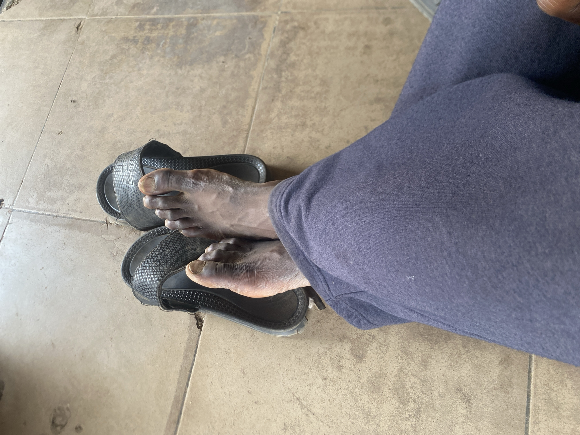 An indoor scene shows a person wearing navy blue sweatpants, sitting with bare feet partially in black sandals on a tiled floor. The tiles are a light brown or tan color with visible grout lines. The person's feet are ashy in color, positioned in the sandals. The setting appears to be in a building in Bukuru, Nigeria.