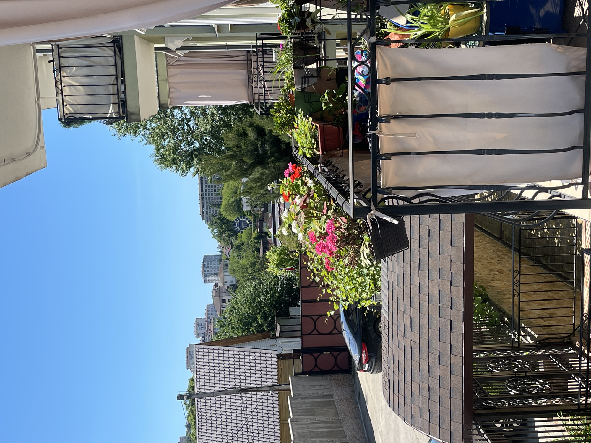 This image captures a vibrant outdoor scene from what appears to be a balcony or terrace, offering a view over a residential area, under a clear blue sky in Sochi, Russia.
In the immediate foreground, to the right, a black metal railing adorned with intricate scrollwork is prominent. White canvas panels are attached to the railing, creating privacy or shade. Numerous potted plants with lush green foliage and brightly colored flowers, including reds and pinks, are arranged along the railing. A dark brown shingled roof of a building below is visible, partially covering a black car parked on a paved surface. To the left, part of a white or light-colored overhang or balcony structure is seen.
The midground features more residential buildings with similar shingled roofs, interspersed with abundant green trees. Another balcony with a light-colored curtain or awning is visible on an adjacent building. A paved path or driveway snakes through this section.
In the background, a clear, expansive blue sky dominates, with several multi-story city buildings and structures visible on the horizon, indicating an urban setting beyond the immediate residential area. The daylight is bright, suggesting a sunny day.
There are no people or visible text in the image. The overall impression is one of a peaceful and well-maintained outdoor living space, characteristic of a pleasant climate.