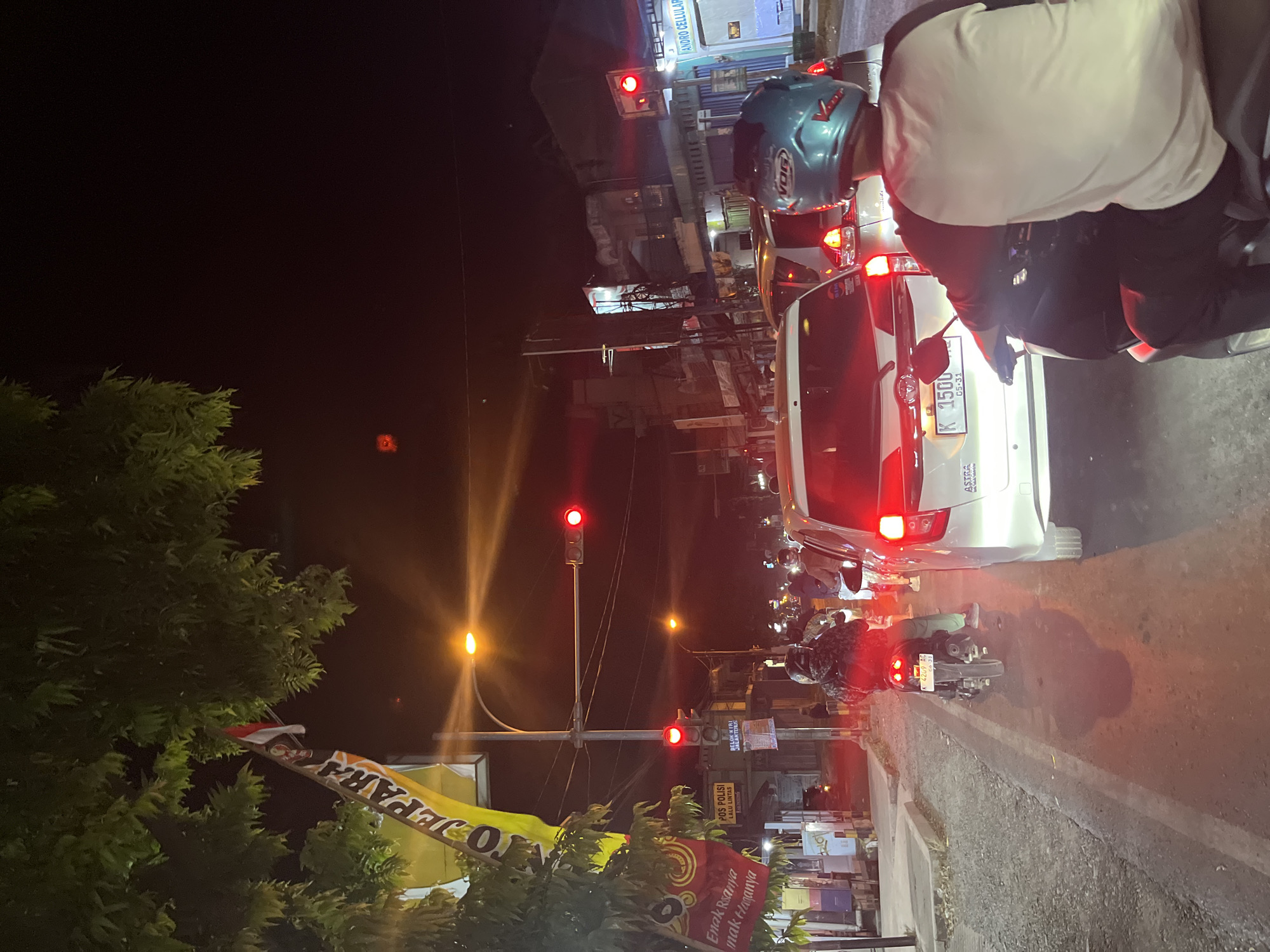 At night, a traffic jam is occurring on a road in Kalinyamatan, Indonesia. A traffic light is red, indicating that vehicles must stop. Several motorcycles and cars are present, with their headlights and taillights illuminating the scene. On the left side of the road, there is a flag with the words "JEPARA" and an Indonesian flag, suggesting the location is near Jepara. A banner below it reads "Enak rasanya, enak rasanya," which translates to "It tastes good, it tastes good." There is also a sign for "POS POLISI" (Police Post) and "BELOK KIRI JALUR BEBAS" (Left Turn Free Lane). The street appears to be lined with shops, some of which are lit. A person wearing a blue helmet is on a motorcycle in the foreground, facing away from the camera, looking at the traffic.