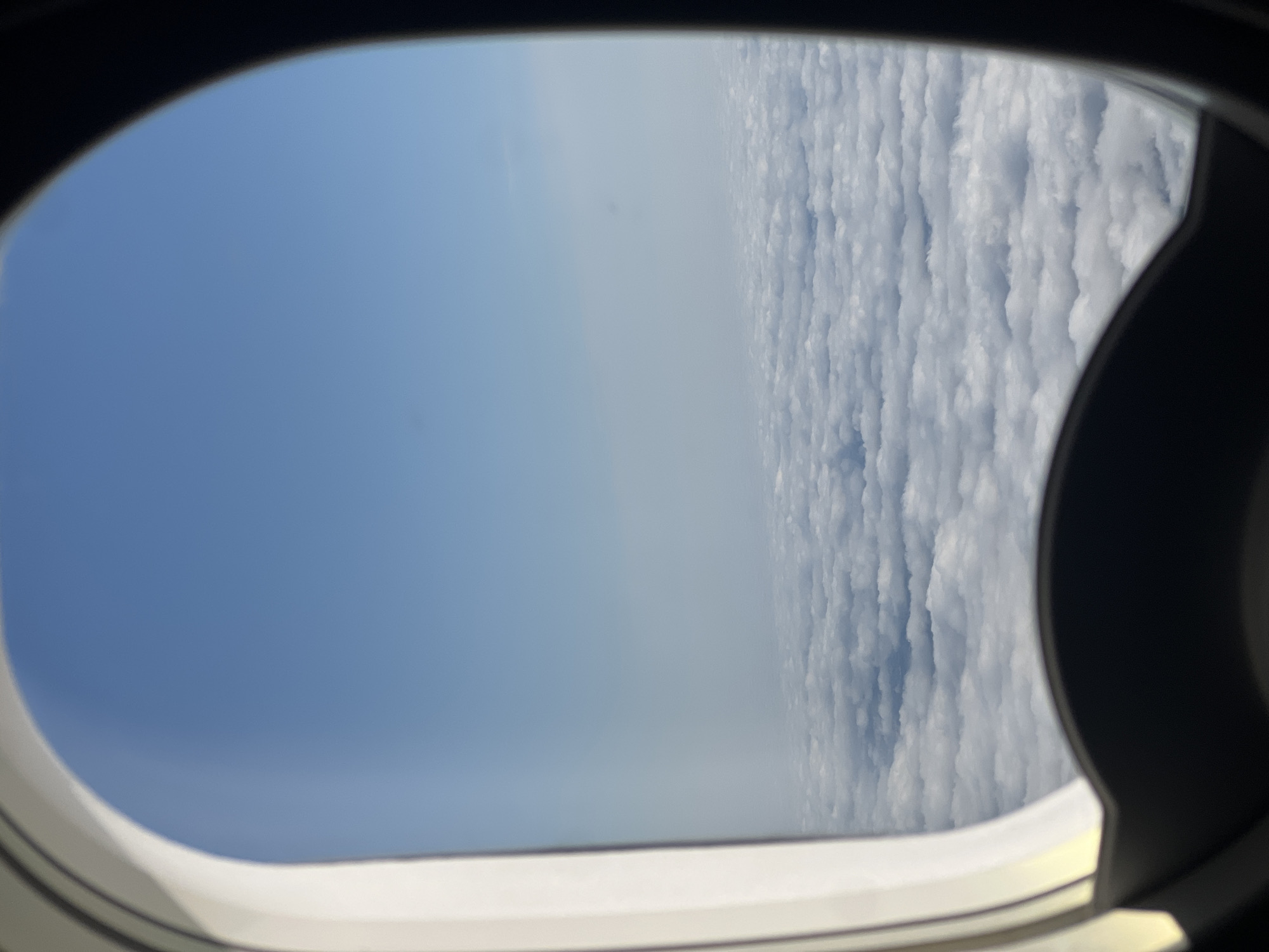 An airplane window frames a view of a vast expanse of white clouds stretching to the horizon under a clear blue sky. The window frame is dark on the right side and white on the left. The clouds appear fluffy and textured, suggesting they are at a significant altitude. No city or specific geographical features are visible, making it impossible to ascertain a location in Kczewo, Poland. The scene depicts a typical view from an airplane in flight.