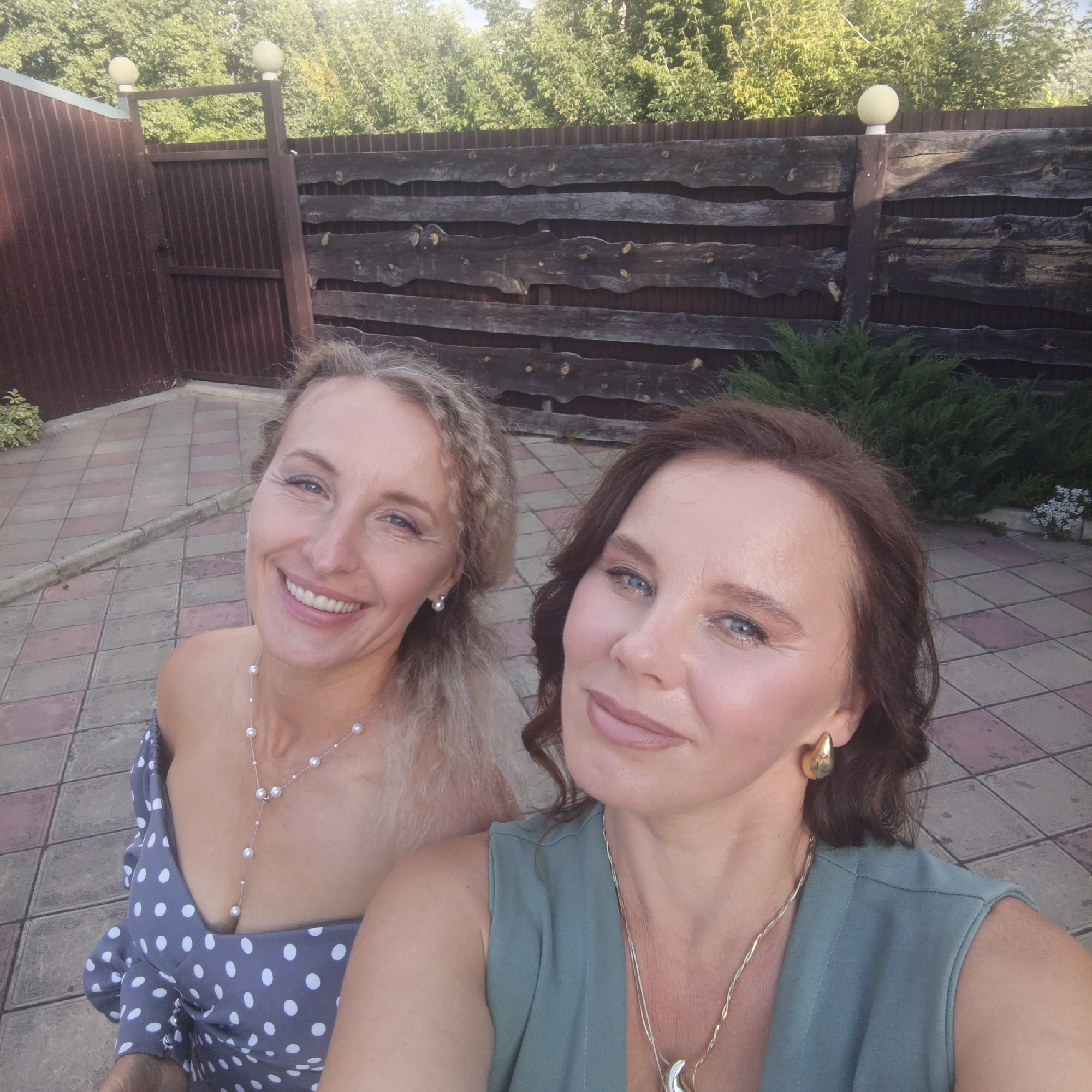 In the city of Chaikovsky, Russia, two women pose for a selfie in an outdoor setting, likely a backyard. The woman on the left has blonde, curly hair and wears a blue and white polka dot dress with a pearl necklace. She is smiling broadly, showing her teeth, and her head is tilted slightly to the right. The woman on the right has dark brown, wavy hair and wears a green sleeveless top with a gold necklace and large gold earrings. Her lips are slightly parted as she looks toward the camera. The background features a rustic wooden fence, a brown metal gate, and lush green trees. The ground is paved with square tiles in a pinkish-gray pattern. The lighting suggests it is daytime, possibly late afternoon, with sunlight casting some shadows.