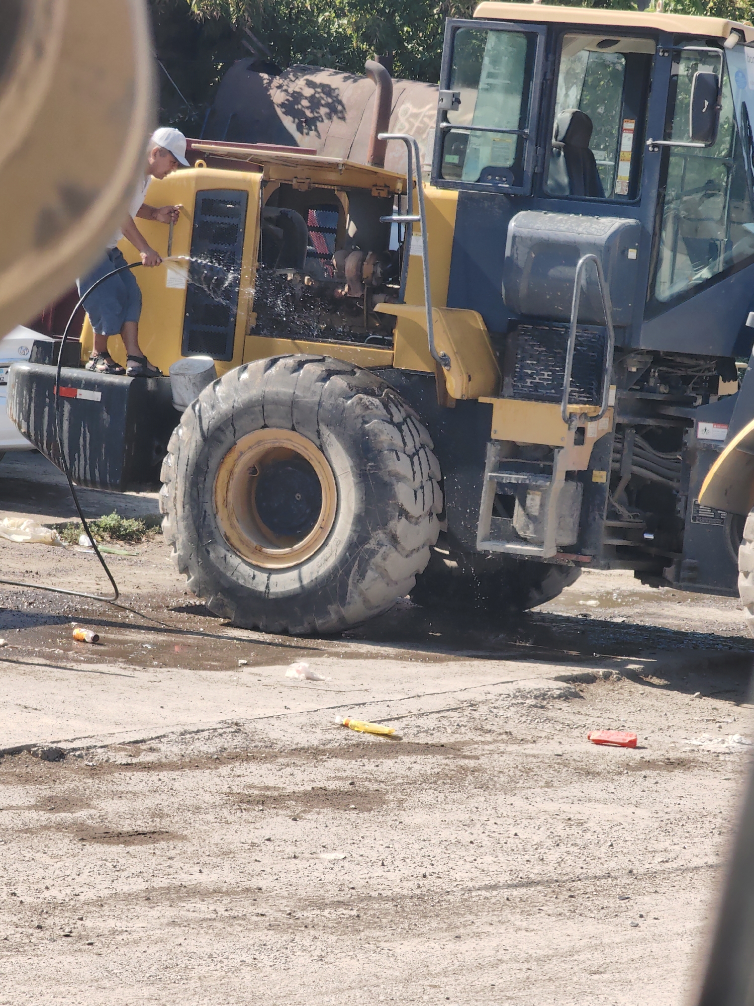 In Shymkent, Kazakhstan, a person wearing a white cap, a white t-shirt, and blue shorts is standing on the side of a large yellow and black piece of construction equipment, possibly a loader, and spraying water with a hose. The construction equipment has a large tire, a cab with tinted windows, and various mechanical components visible. The ground around the equipment is wet and appears to be made of dirt and gravel. There are also some discarded items on the ground, including an orange and white can and a yellow object. The scene is outdoors, with trees visible in the background.