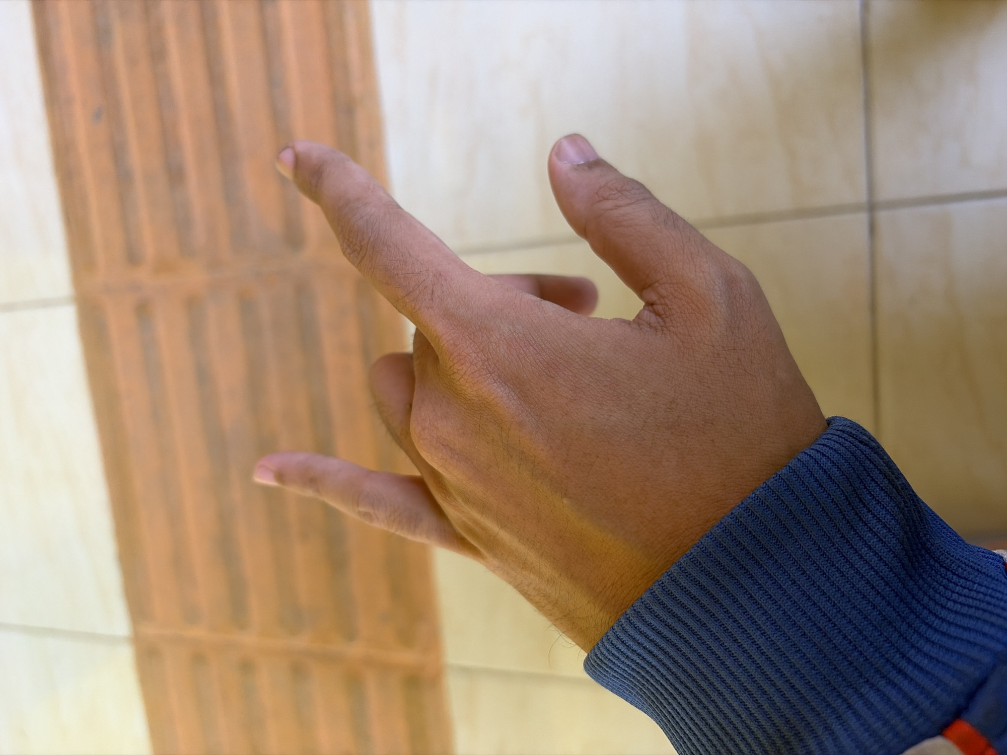 A close-up, overhead shot shows a person's right hand performing the "rock on" or "horns" gesture. The hand is positioned in the center of the frame, with the index finger and pinky finger extended upwards, while the middle and ring fingers are curled downwards and held by the thumb. The hand is wearing a dark blue ribbed cuff, likely from a long-sleeved garment. The background consists of a tiled floor, with a textured, orange section on the left half and a lighter, smoother tiled section on the right. The lighting suggests an indoor environment. This image was captured in Pasekaran, Indonesia.