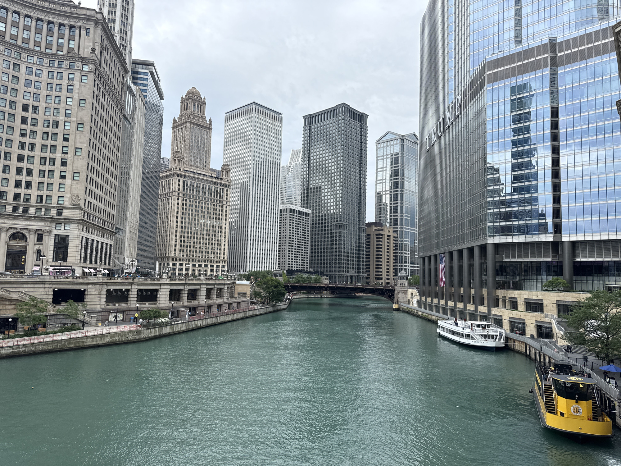 The image depicts a wide, turquoise river flowing through a dense urban landscape, characteristic of Chicago. Tall, modern skyscrapers with glass facades dominate the skyline on either side of the river, reflecting the overcast sky. On the left bank, older, ornate stone buildings stand, featuring classical architectural elements.
Several boats are docked along the right bank. One is a white passenger boat named "WENDELA." Another is a bright yellow vessel with a black upper deck, appearing to be a tour boat, with "CHICAGO" visible on its side. People are seen walking along the riverfront promenade, suggesting a leisurely daytime activity.
The weather appears to be overcast and possibly cool, given the absence of bright sunlight and the appearance of the sky. The overall mood is one of urban grandeur and activity, with the river acting as a central artery through the city. The prominent "TRUMP" logo is visible on the side of one of the glass skyscrapers. The scene captures a typical day on the Chicago River, with its blend of historic and contemporary architecture, and the constant movement of watercraft.
