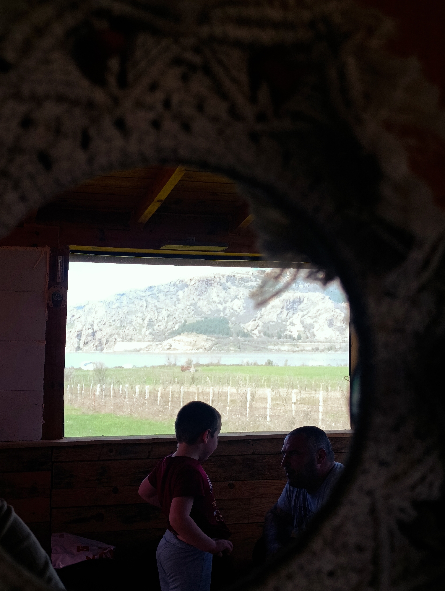 A boy and a man are inside a wooden structure looking out a window at a lake and mountain landscape. A crocheted circular object or frame is in the foreground. Outside the window is a green field and a lake with mountains in the background. The window is within a wooden structure with a yellow-painted beam and a thermometer/gauge hanging on the left frame. The image appears to be taken in Shiroko pole, Bulgaria.