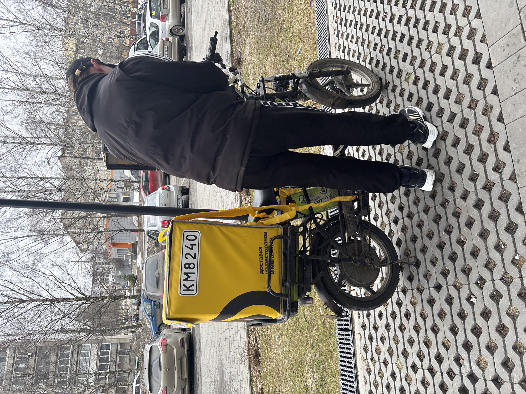 A person wearing a black jacket and black sweatpants with white stripes stands next to a small electric bicycle with a large, yellow delivery box attached to the back. The box has the license plate number "KM8240" and text in Russian that translates to "Delivery from restaurants and stores." The bicycle is parked on a patterned concrete surface with grass and debris visible in the openings. In the background, there are several parked cars, including a silver Renault, a blue car, and other vehicles partially visible. Buildings and bare trees are also visible in the background. The scene appears to be set in a city, possibly Izhevsk, Russia, considering the Cyrillic text on the delivery box and car license plates in the background.