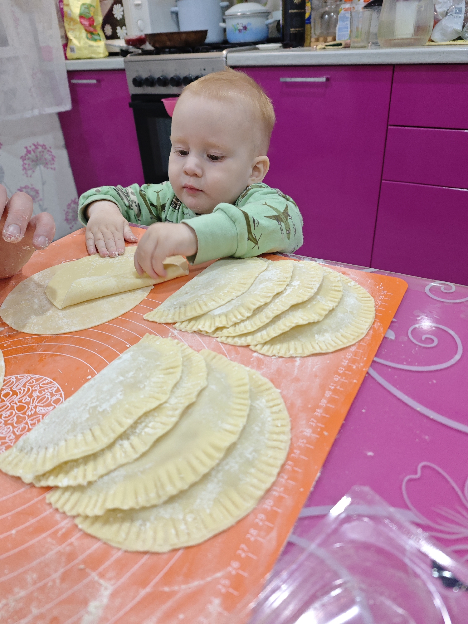 A close-up shot shows a young child in a green patterned shirt at a table. The child is in the process of folding a piece of dough, with several similar dough-filled pastries already prepared nearby. The dough preparation is taking place on orange and pink silicone mats. A hand is visible next to the child, assisting with the cooking. In the background, a kitchen scene unfolds, with hot pink cabinets, a stovetop, and assorted kitchen items visible. The image suggests a cooking activity involving a child, in a home kitchen setting. The location is Markovskii, Russia.