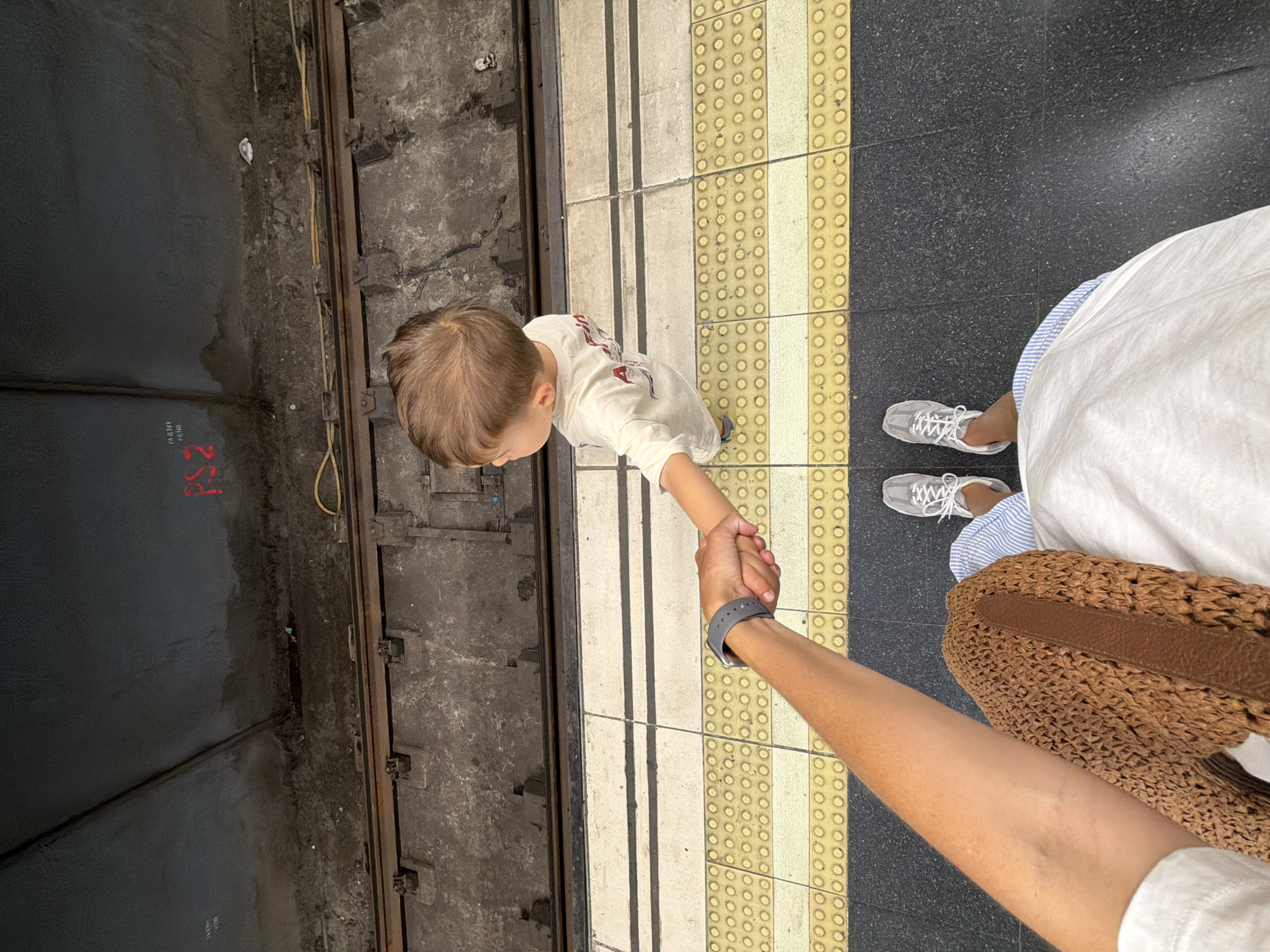 A high-angle, first-person perspective shot shows a person's arm and hand holding the hand of a young child. The individuals are standing on a subway platform in l'Hospitalet de Llobregat, Spain. The platform has a yellow tactile paving strip with raised circles along the edge, bordering a dark grey tiled area. Beyond the platform are subway tracks with wooden sleepers and metal rails, situated within a dark, possibly underground, tunnel structure. The child, facing away from the camera, wears a light-colored long-sleeved shirt. The person's arm displays a grey smartwatch, and a brown woven bag is visible near their leg. The floor of the platform is dark grey speckled material. The tunnel walls are dark and appear weathered, with some red markings visible in the distance.