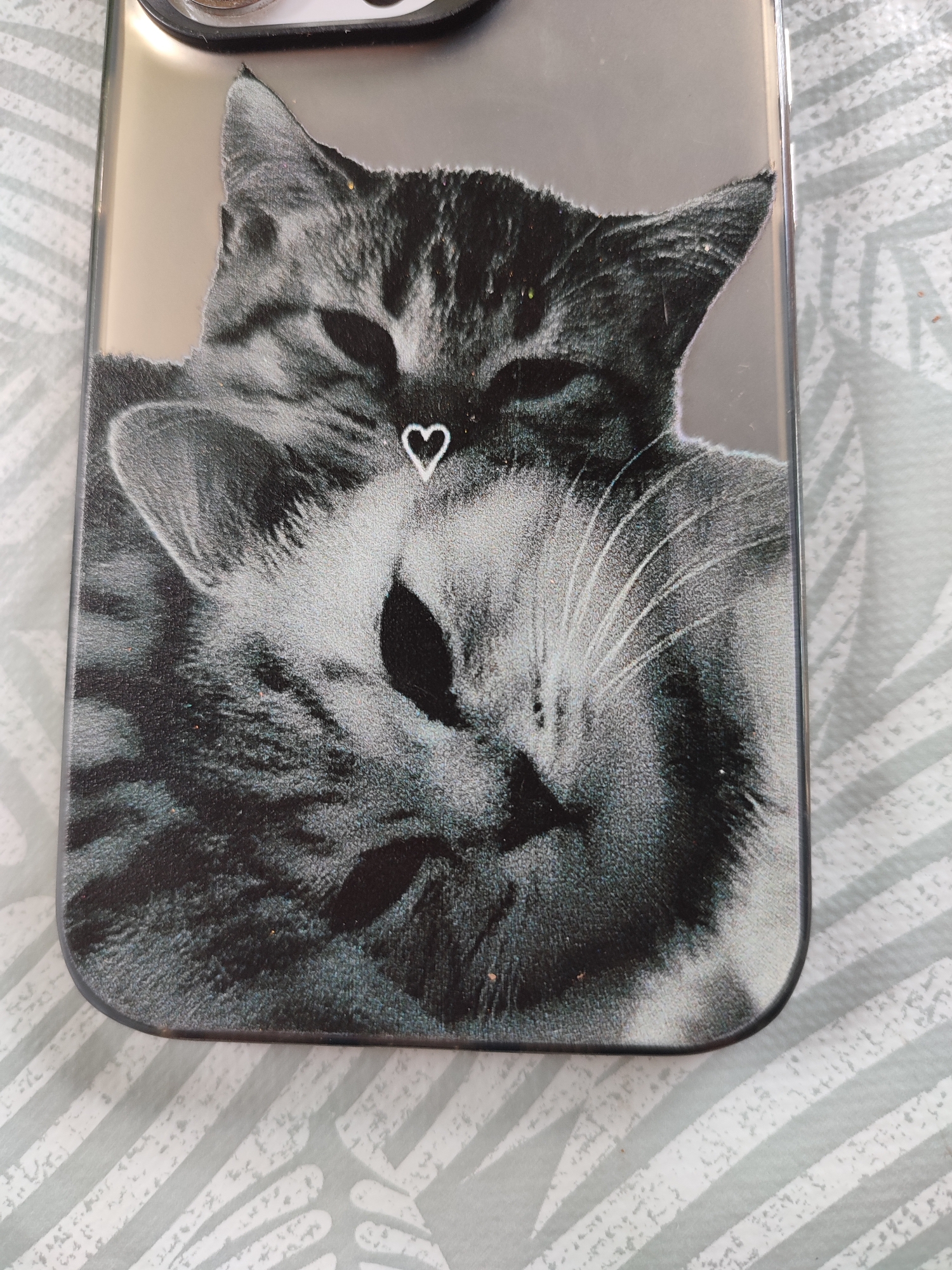 The image shows a close-up of a phone case with a black and white photograph of two cats. The cats are nestled together, with their heads touching, and a small white heart emblem is positioned between their noses. The photo has a vintage, slightly grainy quality. The phone case itself appears to be silver or metallic, and the edges of the phone are visible. The phone case is resting on a surface with a light-colored, textured pattern of what appears to be stylized leaves or palm fronds in shades of grey and white. The overall impression is a sentimental and affectionate depiction of felines.