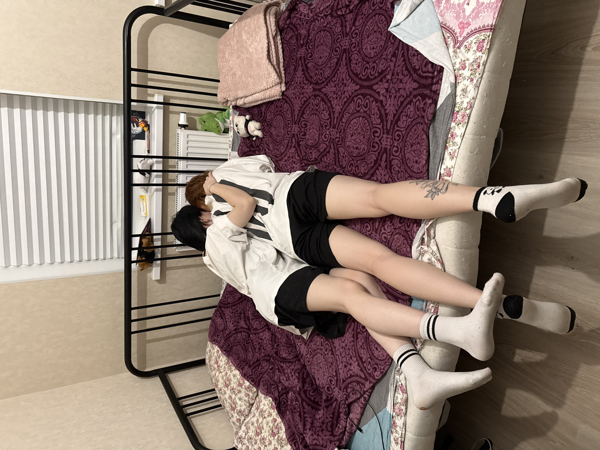 This image depicts a bedroom in Saint Petersburg, Russia, showing two individuals lying embraced on a bed. The bed features a black metal headboard and frame. The mattress is covered by a floral-patterned sheet, a purple blanket with a classical design, and a light blue/grey blanket on the left. A folded, light pink textured blanket rests on the right side of the bed.
The individual on the left has dark hair, wears a plain white short-sleeved t-shirt, black shorts, and white socks with two black stripes. The individual on the right has reddish-brown hair, wears a white short-sleeved t-shirt with black vertical stripes on the back, black shorts, and white socks with a black design (possibly a panda face) on the heel. A tattoo, possibly floral or leafy, is visible on the lower right leg of this individual. Their right arm is wrapped around the other person's upper body. Both individuals are lying prone, facing away from the camera, with their heads close together in an intimate embrace.
The room has light beige, textured wallpaper. A window with horizontal white Venetian blinds is visible at the top right, with a white shelf mounted below it. A white panel radiator is situated below the shelf. Various small objects, including a yellow box, a white plush item, and a small brown plush animal, are on the shelf. A green alligator plush toy is positioned on the bed near the headboard, and a small white plush cat with a black collar lies on the purple blanket further down the bed. The floor is light-colored wood or laminate, and a black cable is visible on the left side of the bed. The lighting is artificial, creating some shadows.