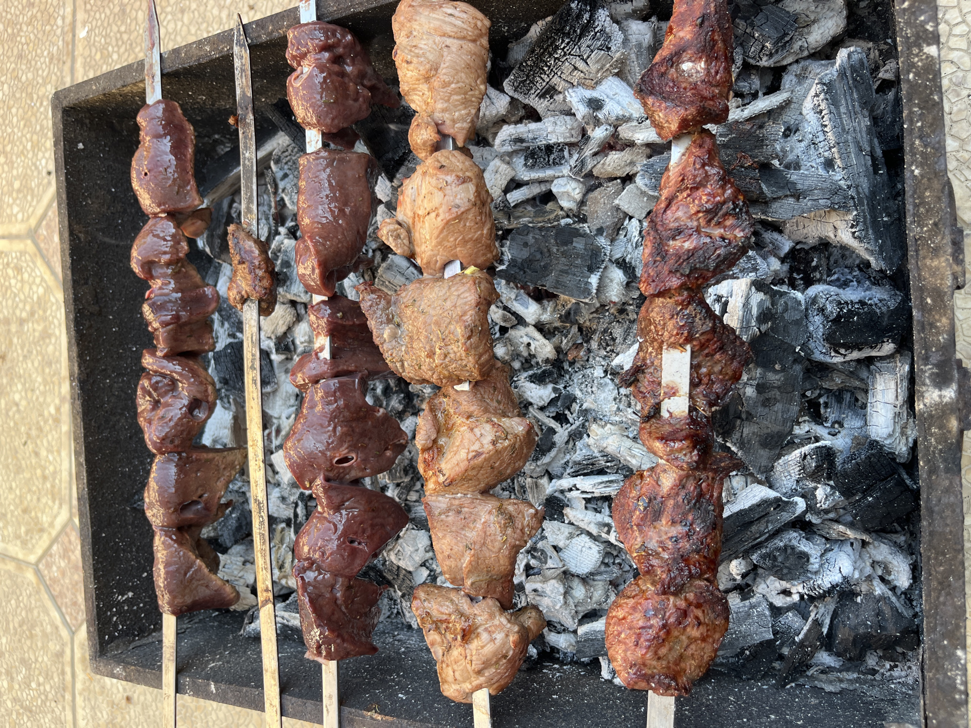 The image shows three metal skewers placed over hot coals in a rectangular grill. The left skewer holds what appears to be liver, the middle skewer has pieces of meat that are lighter in color, and the right skewer has darker, grilled meat. The coals beneath are a mix of black and ash-covered pieces. The location is Kaspiysk, Russia.