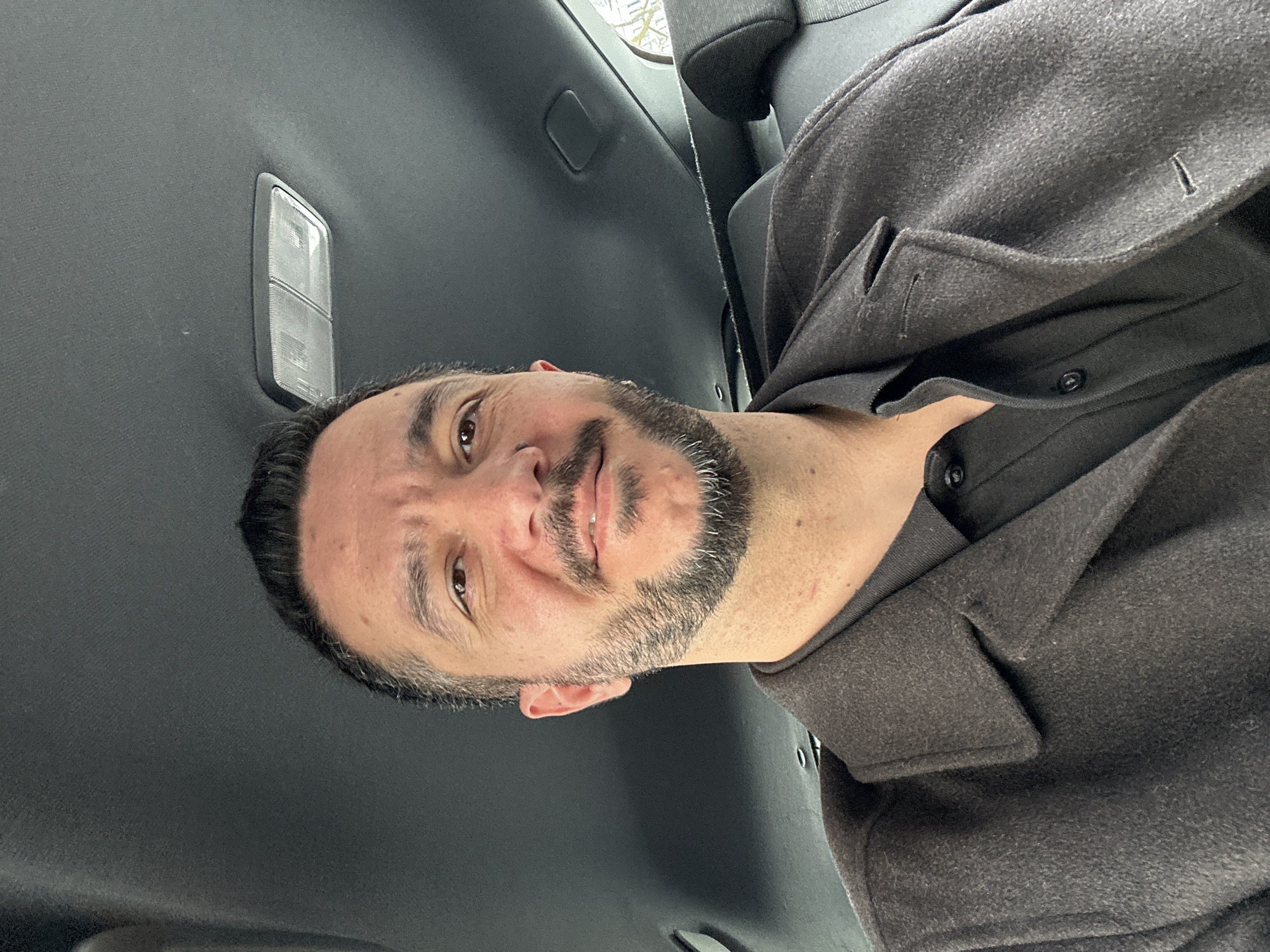 A man with short, dark hair and a salt-and-pepper beard is sitting in a car, possibly in the back seat. He is wearing a dark-colored coat over a shirt with visible buttons. The interior of the car is primarily dark gray or black, with a dome light visible on the car's ceiling. The location is not discernible from the visual information provided.