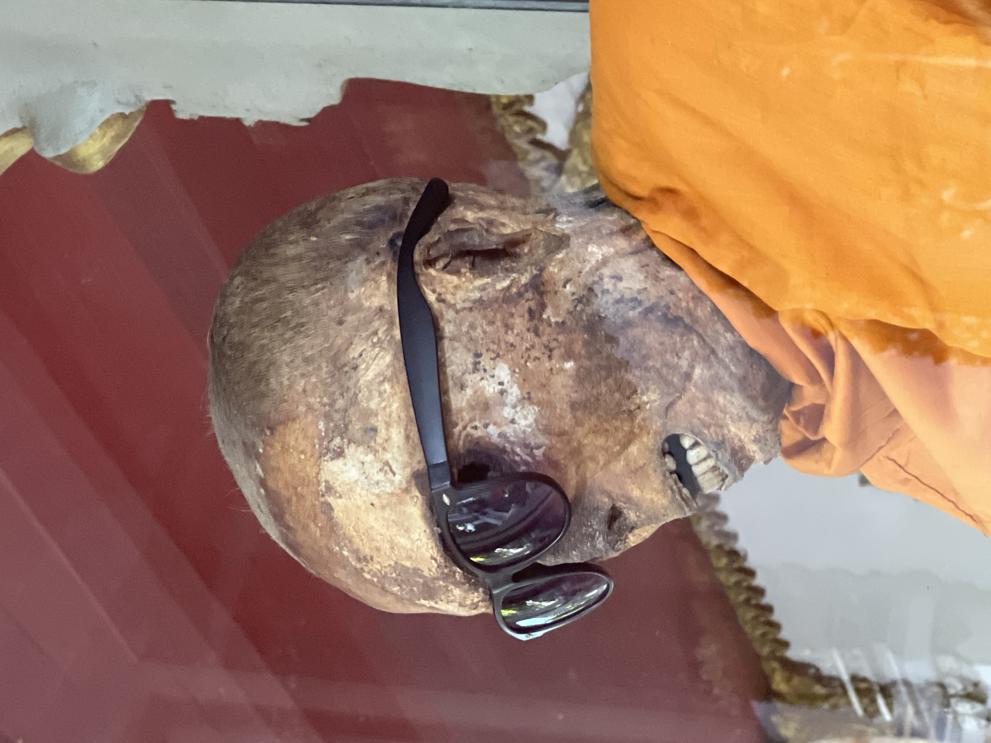 In Baan Hua Thanon, Thailand, a mummified monk is displayed in a glass case, wearing sunglasses and an orange robe. The monk's skin is leathery and discolored, with visible teeth and bone structure. The background of the display includes red and gold decorative elements.