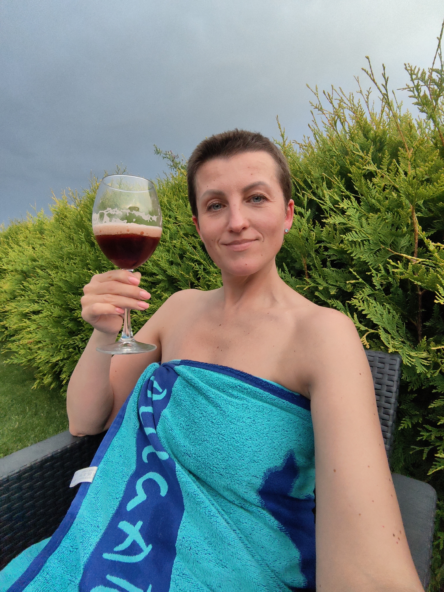 A woman with short brown hair, wearing a teal and blue striped towel, holds a glass of dark red liquid in her right hand. She is seated in a dark outdoor chair, with a manicured green lawn to her left and a dense green hedge behind her. The sky above is overcast and gray. The location is the city of Fanipalski sel'ski Savet, Belarus.