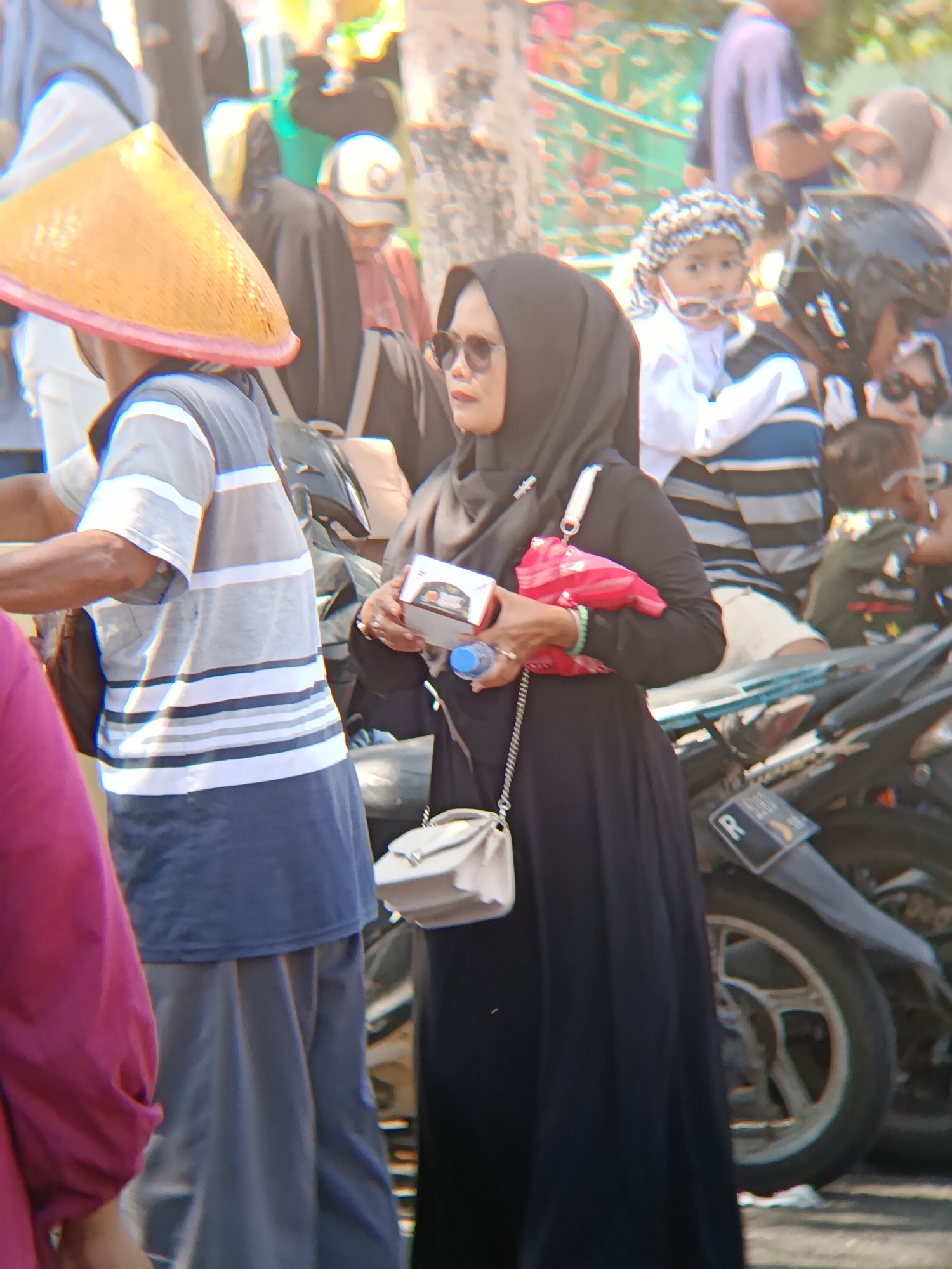 The image depicts a street scene in Desa Kebon Manis, Kecamatan Cilacap Utara, Indonesia, with several individuals visible amidst parked motorcycles and an outdoor environment suggesting a public gathering or market. In the foreground, a person wearing a black hijab and sunglasses is holding a small box and a blue water bottle, with a red fabric tucked into their arm and a light-colored purse slung over their shoulder. To the left, another individual is partially visible, wearing a conical hat and a striped shirt, carrying a dark bag. Behind them, a child wearing a white shirt and a patterned head covering is looking forward, positioned between adults. The background shows a blur of other people and a motorcycle. The overall lighting suggests a bright day.