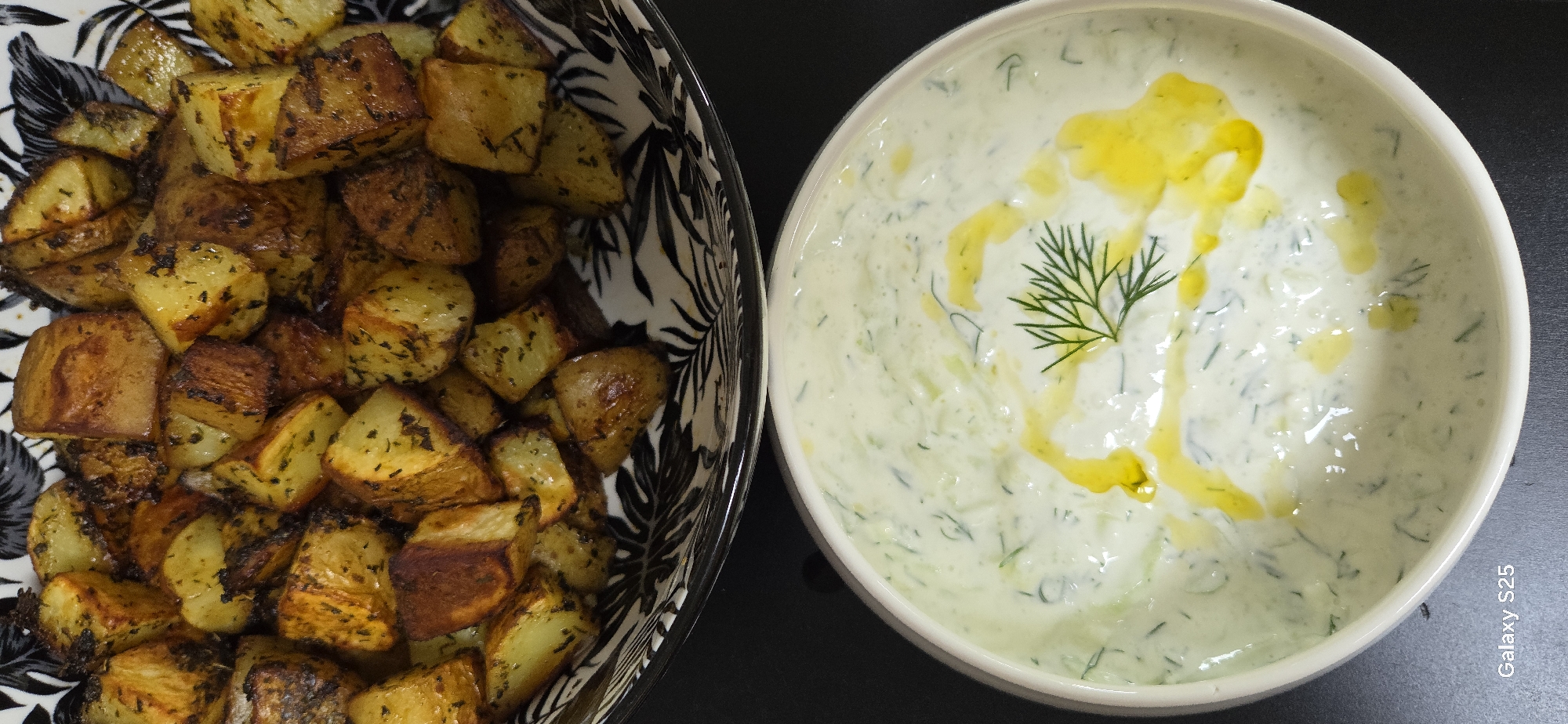 The image shows a close-up view of two food dishes. In the upper portion of the image, a bowl contains roasted potato wedges seasoned with herbs, appearing crispy and golden brown. The bowl has a decorative black and white pattern around its rim. Below this, a white bowl holds a creamy white dip, possibly yogurt-based, with finely chopped green herbs and shredded cucumber. Drizzled over the dip are droplets of yellow oil, and a sprig of fresh dill is placed on top as a garnish. The dishes are set against a dark, flat surface. The bottom left corner of the image displays the text "Galaxy S25". There is no discernible information in the image to suggest a location in Eeklo, Belgium.