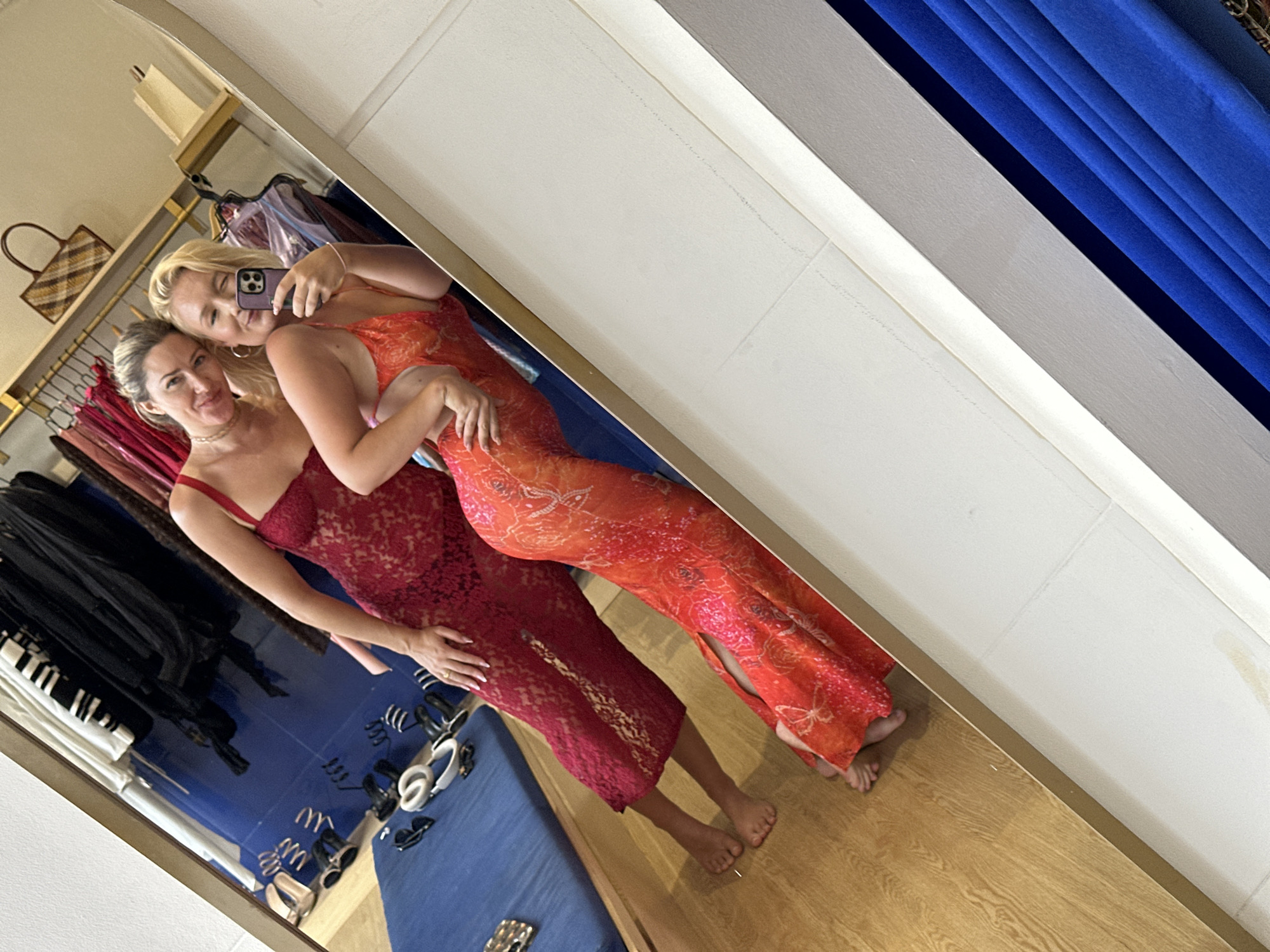 Two Caucasian women are posing for a selfie in front of a mirror. The woman on the left has blonde hair pulled back and is wearing a red lace dress. The woman on the right has blonde hair and is wearing an orange floral dress. They are in a clothing store, likely in North Kuta, Indonesia, based on the environment and the type of clothing. There are clothes hanging on a rack behind them, and a blue curtain to the right of the mirror. A bag is visible on a shelf behind the rack.