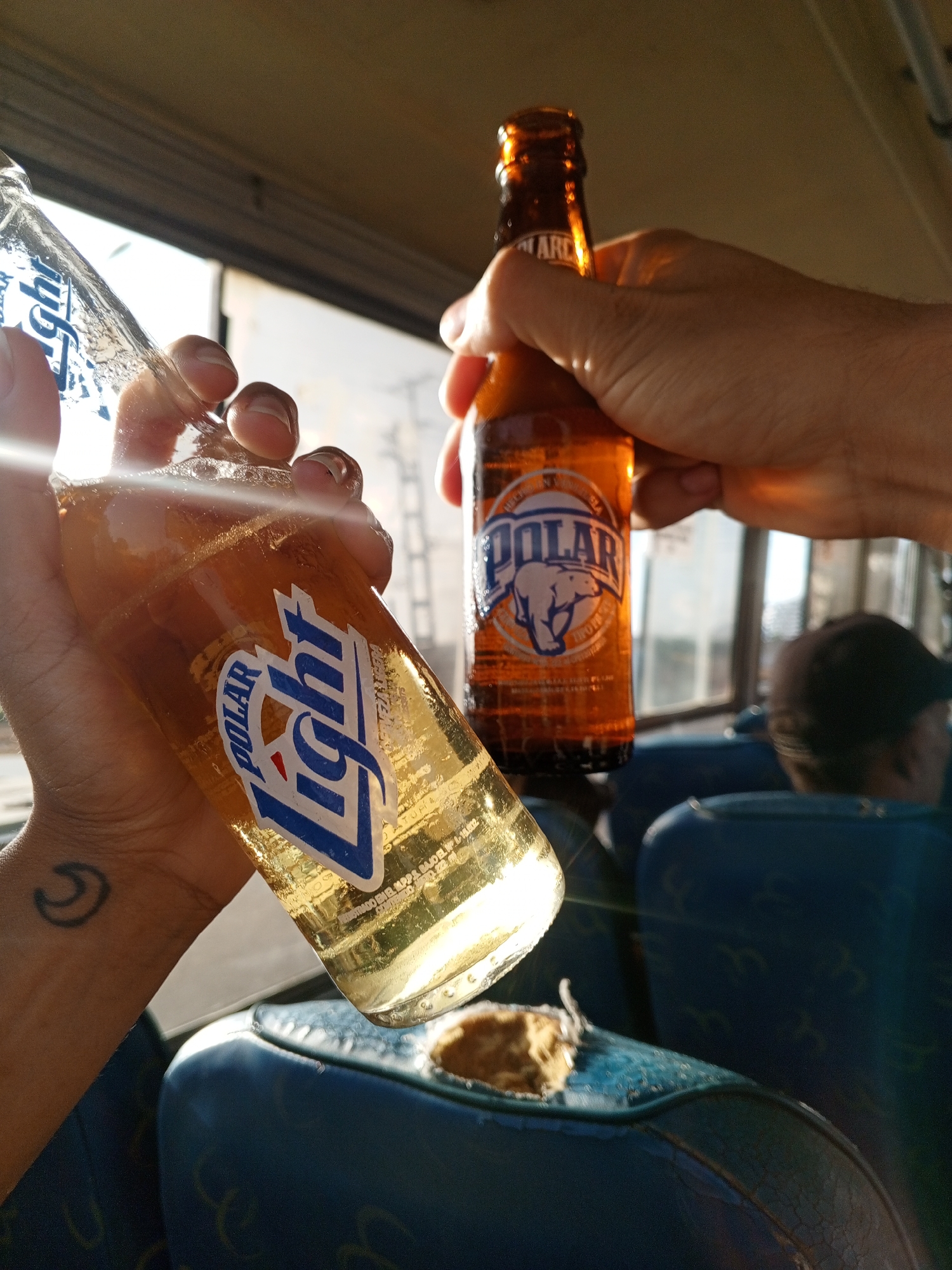 Two hands hold bottles of Polar Light beer. One hand holds a bottle of Polar beer. The bottles are held inside a bus. A man wearing a baseball cap sits in a seat. A seat has a hole in it. The location could be Porlamar, Venezuela.