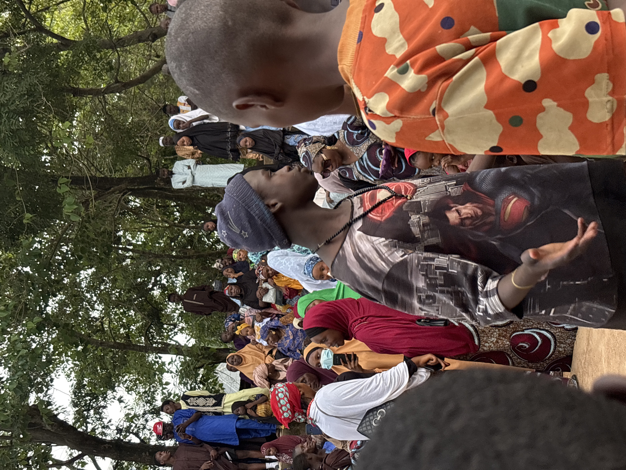 The image depicts a gathering of people outdoors in what appears to be a community event in the city of Jos, Nigeria. The setting is under a canopy of trees with dappled sunlight. A diverse group of individuals, predominantly dressed in traditional African attire and Islamic modest wear, are seen interacting and observing. In the foreground, two young males are facing each other, with one wearing a t-shirt featuring Superman and the other a vibrant orange and white patterned garment. Behind them, a woman in a red headscarf and a light blue face mask is visible, holding a smartphone. Further back, a crowd is seated and standing, many with women and children present. The atmosphere suggests a social or religious occasion.