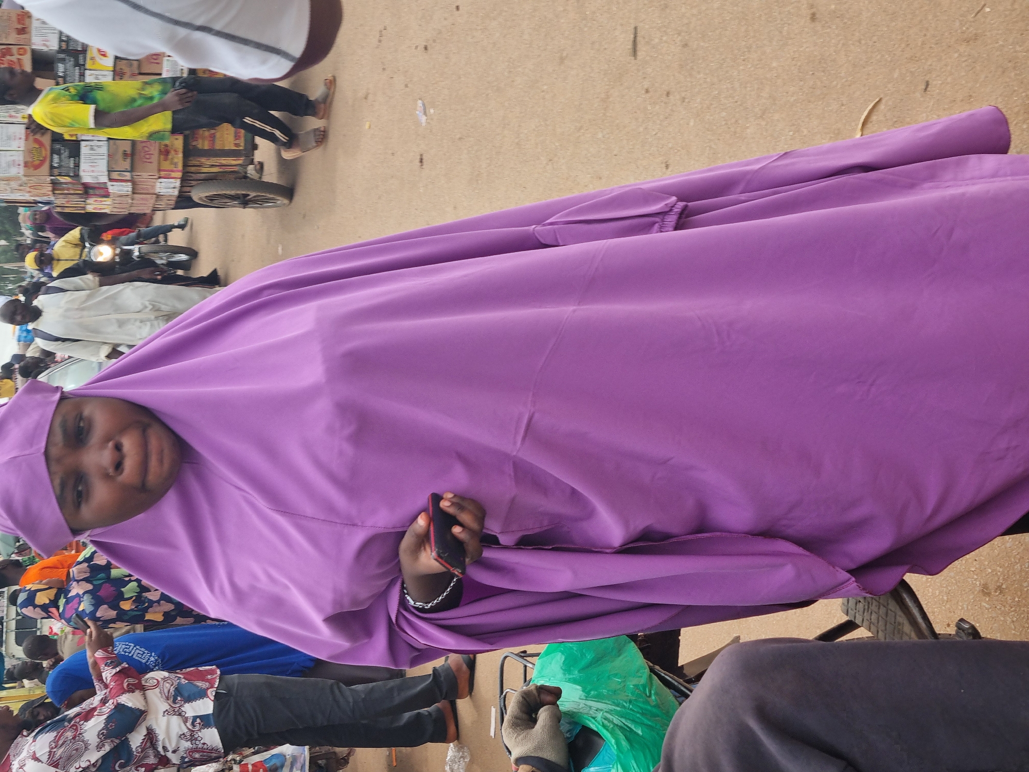 A woman wearing a purple hijab and holding a black cell phone looks toward the camera, likely in Jos, Nigeria. Her hijab covers much of her body as she is sitting on some sort of bicycle, possibly a tricycle. In the background, there are several people, a motorcycle, a cart carrying boxes, and a street.