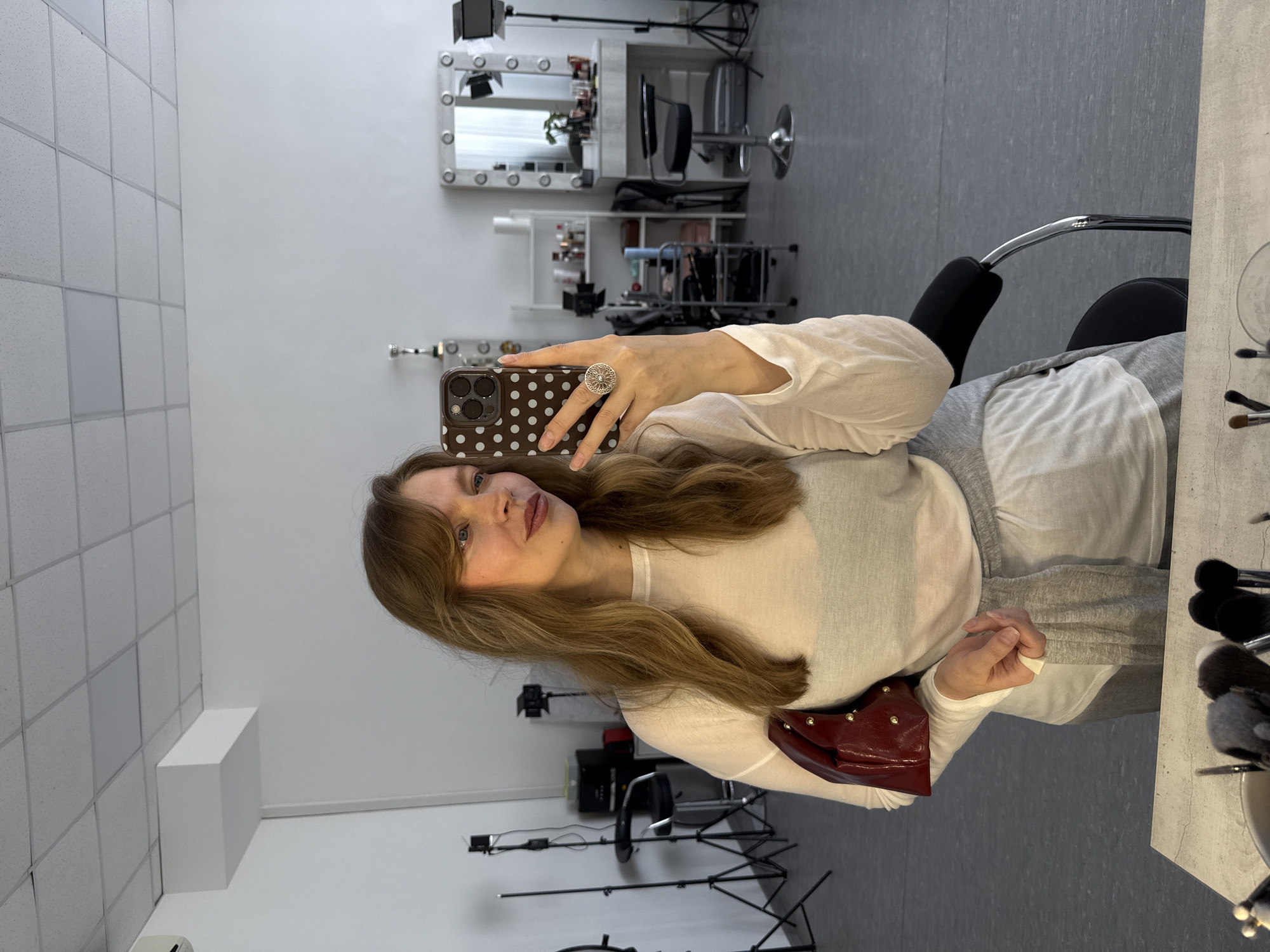 A young woman is captured in a mirror selfie within what appears to be a beauty salon or photography studio in Izhevsk, Russia. She has long, light brown hair with bangs, light blue eyes, and a soft, confident expression with a subtle smile. She is wearing a light-colored, long-sleeved top and grey pants or skirt, with a red, studded handbag resting on her lap. On her right index finger, she wears a prominent silver ring with an intricate, floral-like design. She holds a smartphone with a dark brown, white polka-dotted case, featuring a multi-lens camera module, positioned to take the photo.
The background reveals typical studio elements. To her right, a makeup vanity mirror with exposed Hollywood-style light bulbs is visible, adorned with various cosmetic products and brushes. A black, high-backed swivel chair with a chrome base sits before the vanity. Further back, studio lighting equipment, including tripods and a softbox or light fixture, is set up. The room features white walls, a grey tiled floor, and a grid-patterned acoustic ceiling. The overall scene suggests a professional and functional environment.