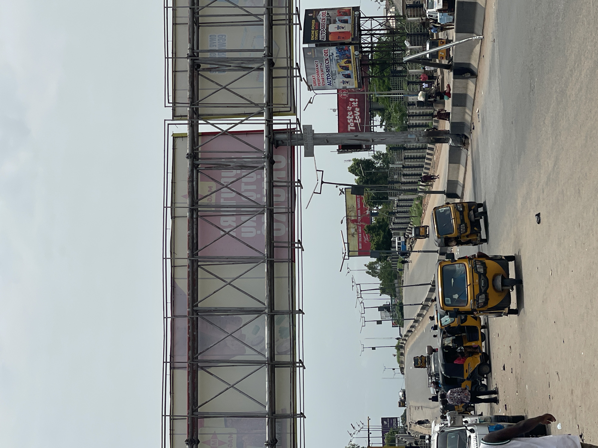 The scene depicts a road in Nkpor, Nigeria. Several yellow tricycles (auto rickshaws) are traveling on the road. A large billboard structure dominates the left side of the frame. Various advertisements, including those for "AUTO-SHECK OIL" and "Taste it... Love it!", are visible on the billboards along the road. There are pedestrians along the road. Streetlights are erected along the road.