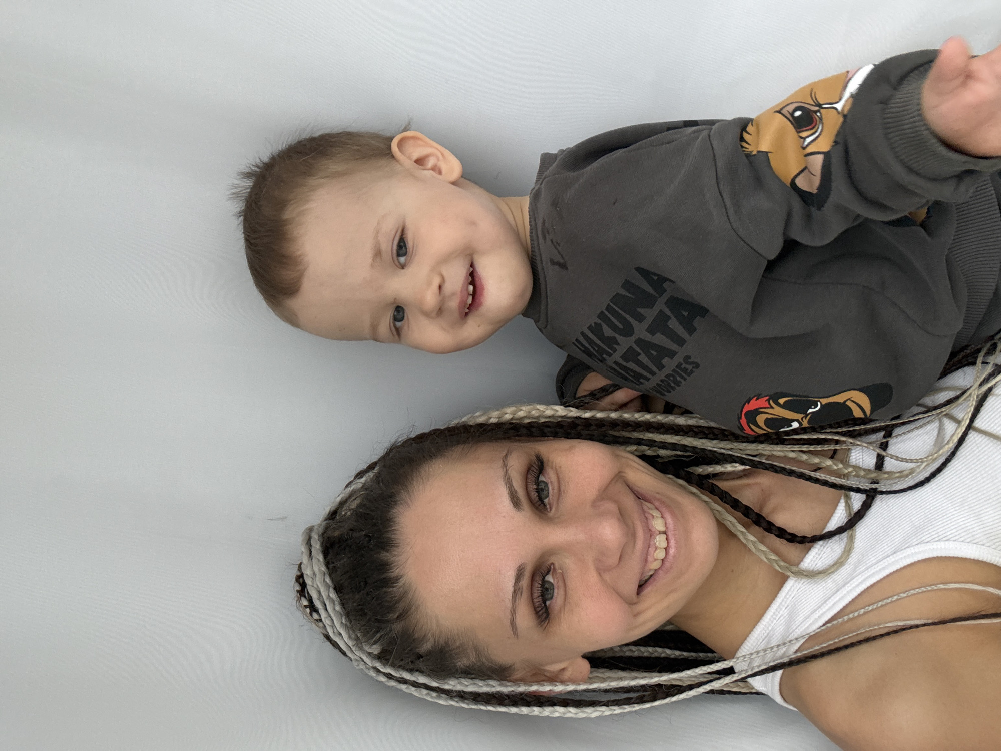 A cheerful portrait features a woman and a young child, both smiling brightly and looking towards the camera, set against a plain light grey or white background. The image appears to be rotated 90 degrees clockwise, suggesting the subjects are lying down or the photo was taken at an angle.
The woman, positioned in the foreground, has an open and friendly expression. She sports distinctive long, multi-colored braids incorporating shades of dark brown, light brown, and white/grey. Her eyes are bright, and her wide smile reveals her teeth. She is wearing a simple white ribbed tank top. Notable details on her face include long, well-defined eyelashes.
Above and slightly behind the woman is a young child, likely a toddler, with light brown hair. The child has striking blue eyes and a wide, joyful smile that shows small bottom teeth and creates charming dimples. The child is dressed in a dark grey long-sleeved sweatshirt. Visible text on the sweatshirt reads "HAKUNA MATATA," with "NO WORRIES" partially visible underneath. Cartoon animal characters, resembling Pumbaa from Disney's The Lion King, are printed on the sleeves and possibly other parts of the garment.
Both subjects convey a sense of happiness and affection, suggesting a warm family moment captured in an informal indoor setting. The plain background focuses attention entirely on their joyful expressions. The photo's context is Rusanovo, Russia.