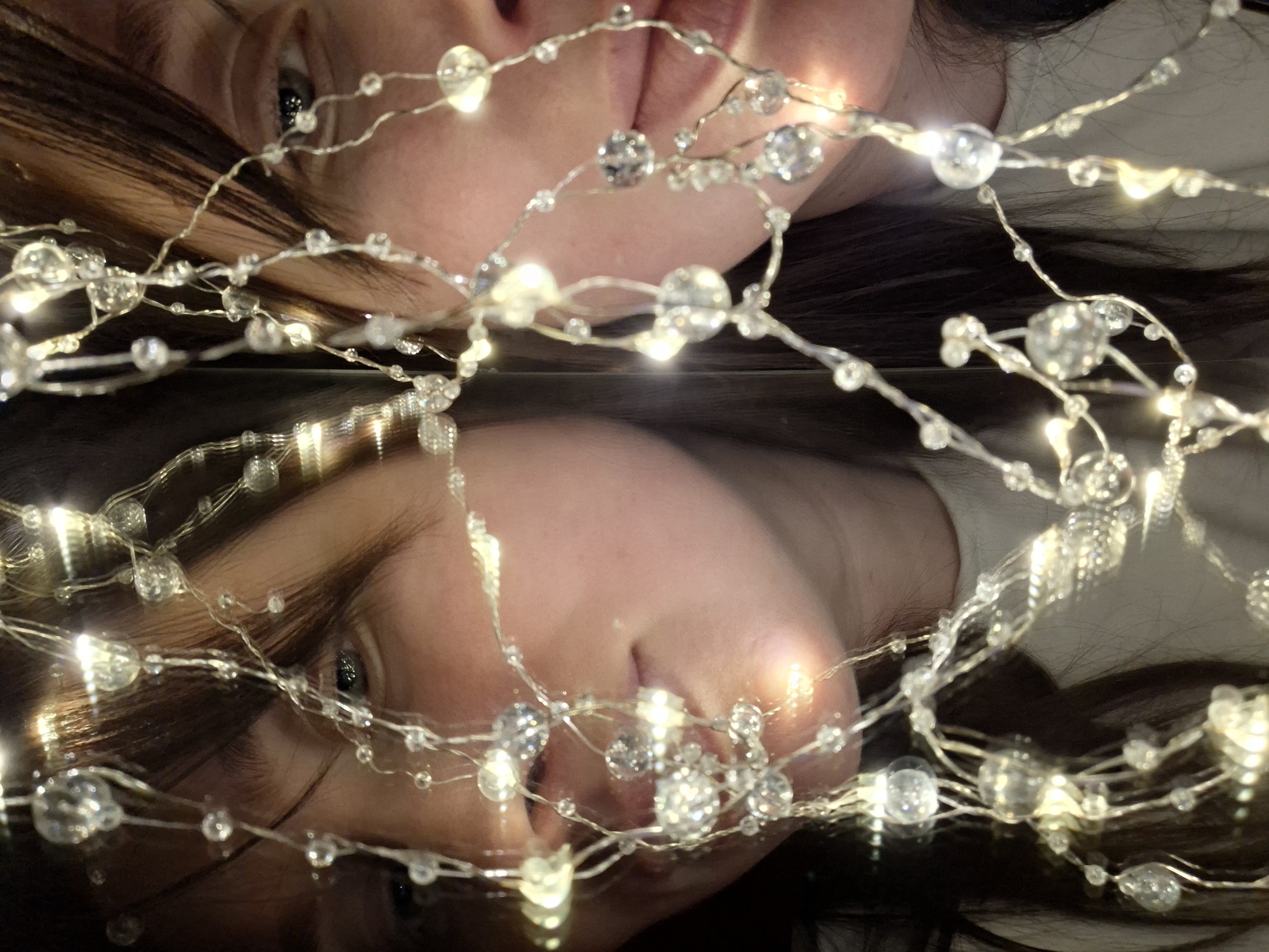 The image shows a close-up, inverted reflection of a person's face on a reflective surface. The upper portion of the image displays the face with features including lips, a nose, an eye, and brown hair. A string of decorative lights with small, round, reflective beads is draped across the face. The lower portion of the image mirrors the face and lights, creating a symmetrical composition. The reflective surface adds a dark, glossy texture to the image. The location and specific details about the city are unknown.
