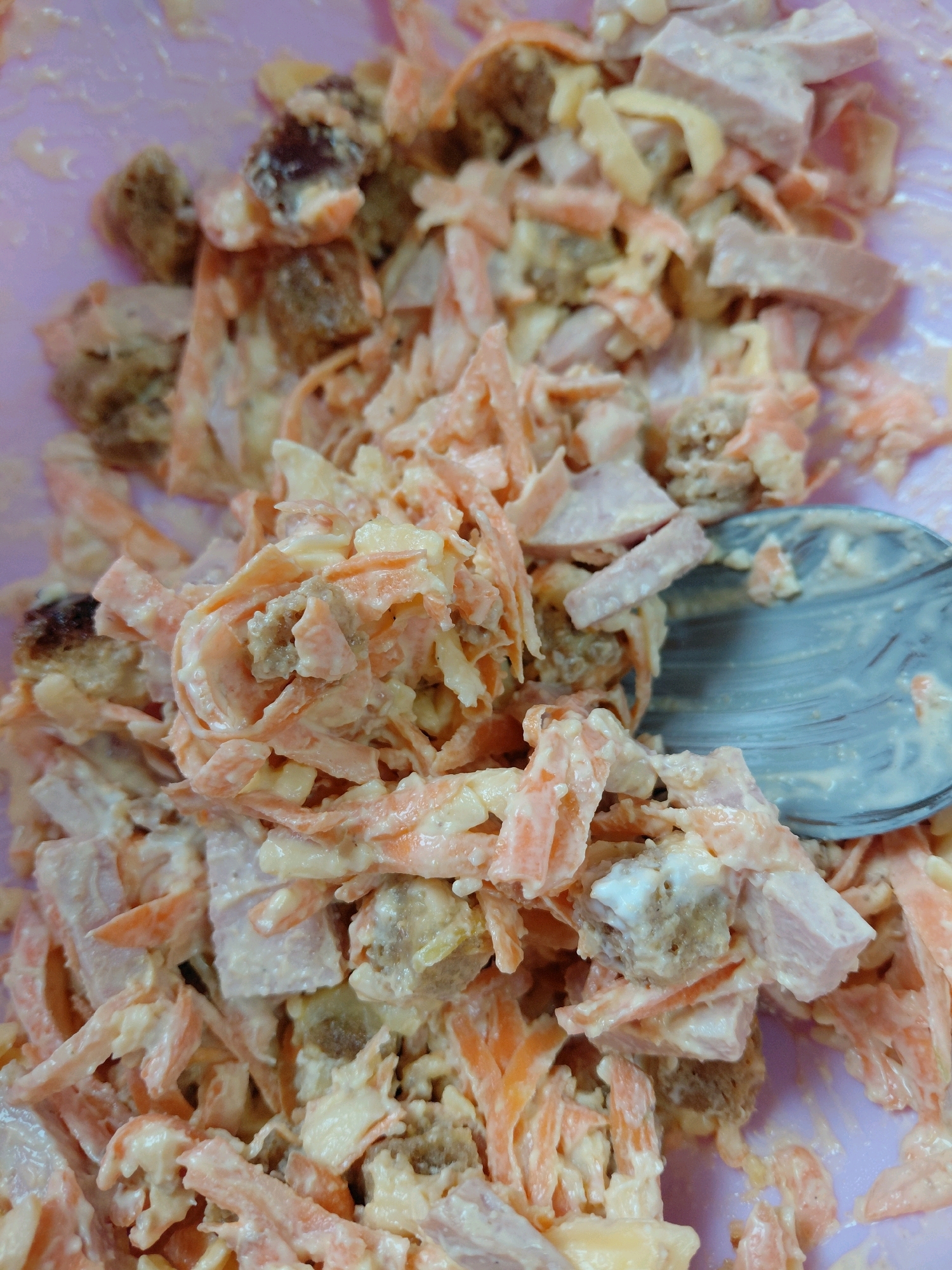 The image displays a close-up, overhead view of a bowl filled with a mixed salad. The salad appears to contain shredded carrots, cubed ham, and small, irregularly shaped pieces of bread or croutons, all coated in a creamy, light-colored dressing. A silver spoon is partially visible on the right side of the frame, resting within the salad and appearing to have some of the mixture on its surface. The bowl itself is a light purple color. The background is out of focus but suggests a home or indoor environment, possibly a kitchen. There are no people visible in the image, and no specific landmarks or indications of the city of Malinovka, Russia, are present. The lighting is even, suggesting a well-lit room.