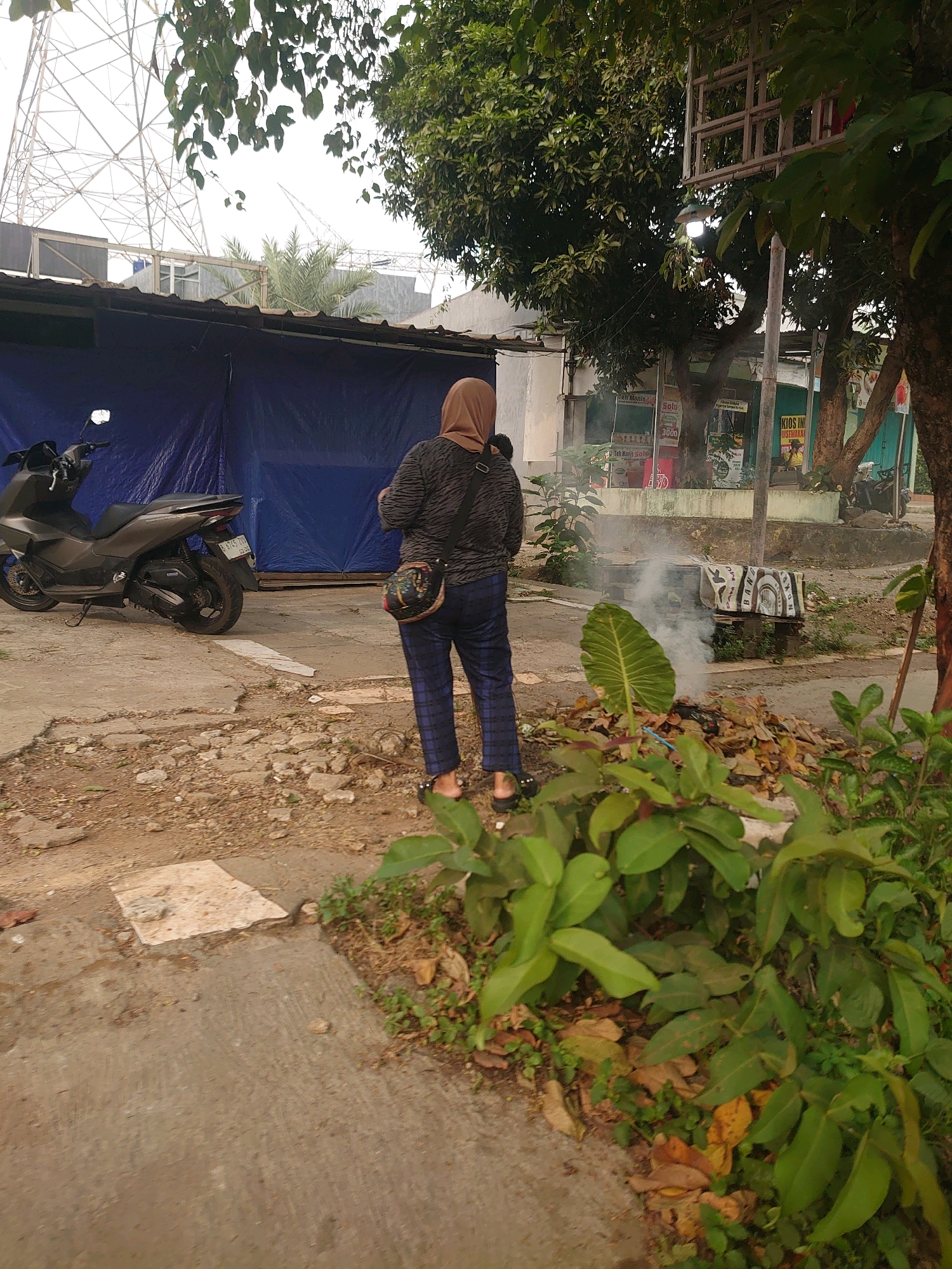 In Depok, Indonesia, a person wearing a brown hijab, a black and white striped shirt, and checkered pants stands in front of a pile of burning leaves that is emitting smoke. A dark gray scooter is parked to the left of the frame, and a blue tarp is visible behind it. The scene appears to be on an outdoor paved area with some debris and vegetation present. In the background, trees, buildings, and a large metal tower structure can be seen.