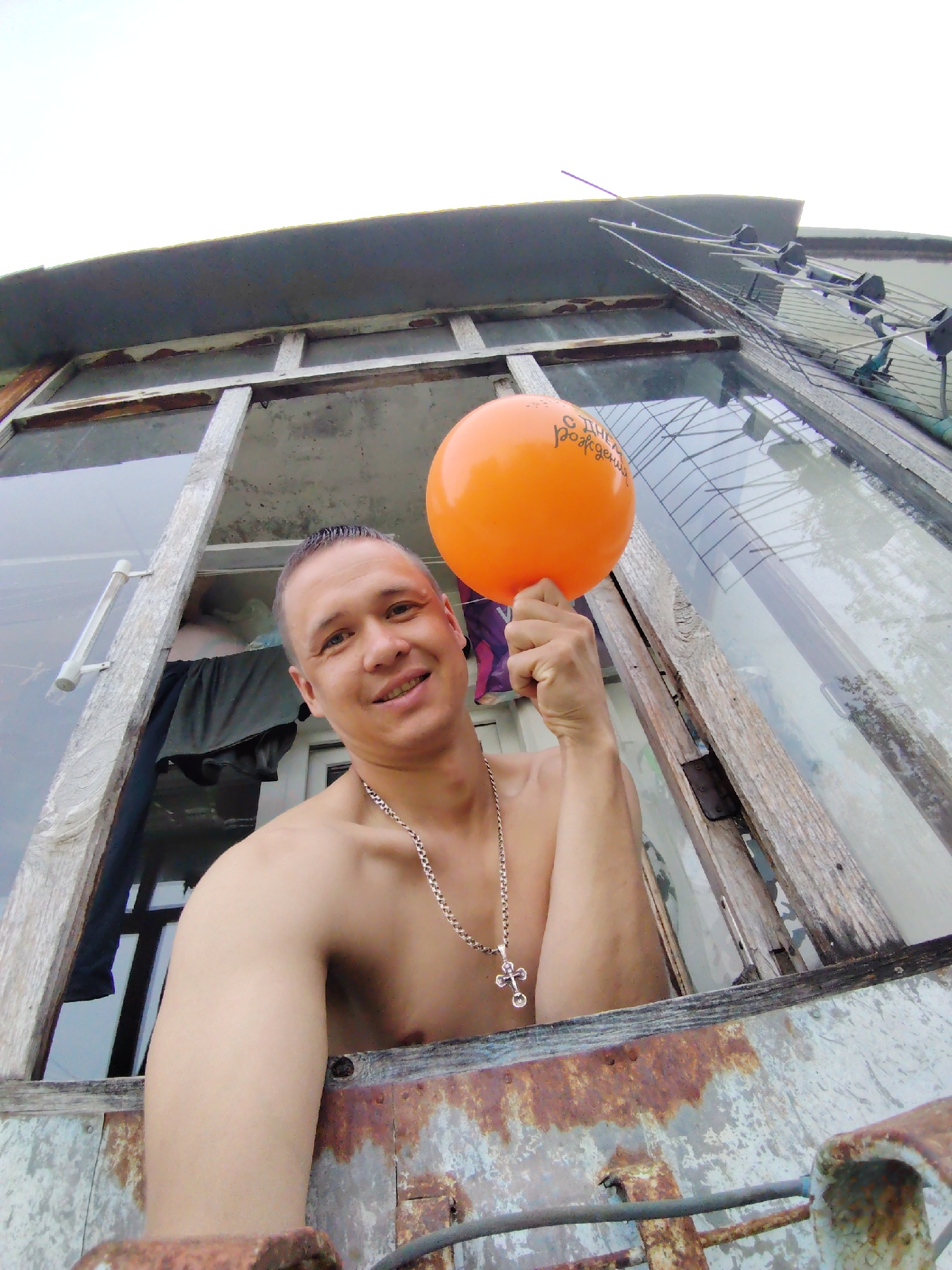 A medium shot shows a shirtless man holding an orange balloon with Cyrillic script. The man has short dark hair and wears a silver cross necklace. He is smiling and appears to be looking at the camera. The man is positioned in the lower left corner of an open, weathered window frame. The frame is rusted and peeling, suggesting it is part of an older structure. Through the window, there are reflections of the outdoors and items inside the structure, including clothing hanging inside. An antenna array is visible above the window. The picture appears to have been taken in Chaikovsky, Russia.