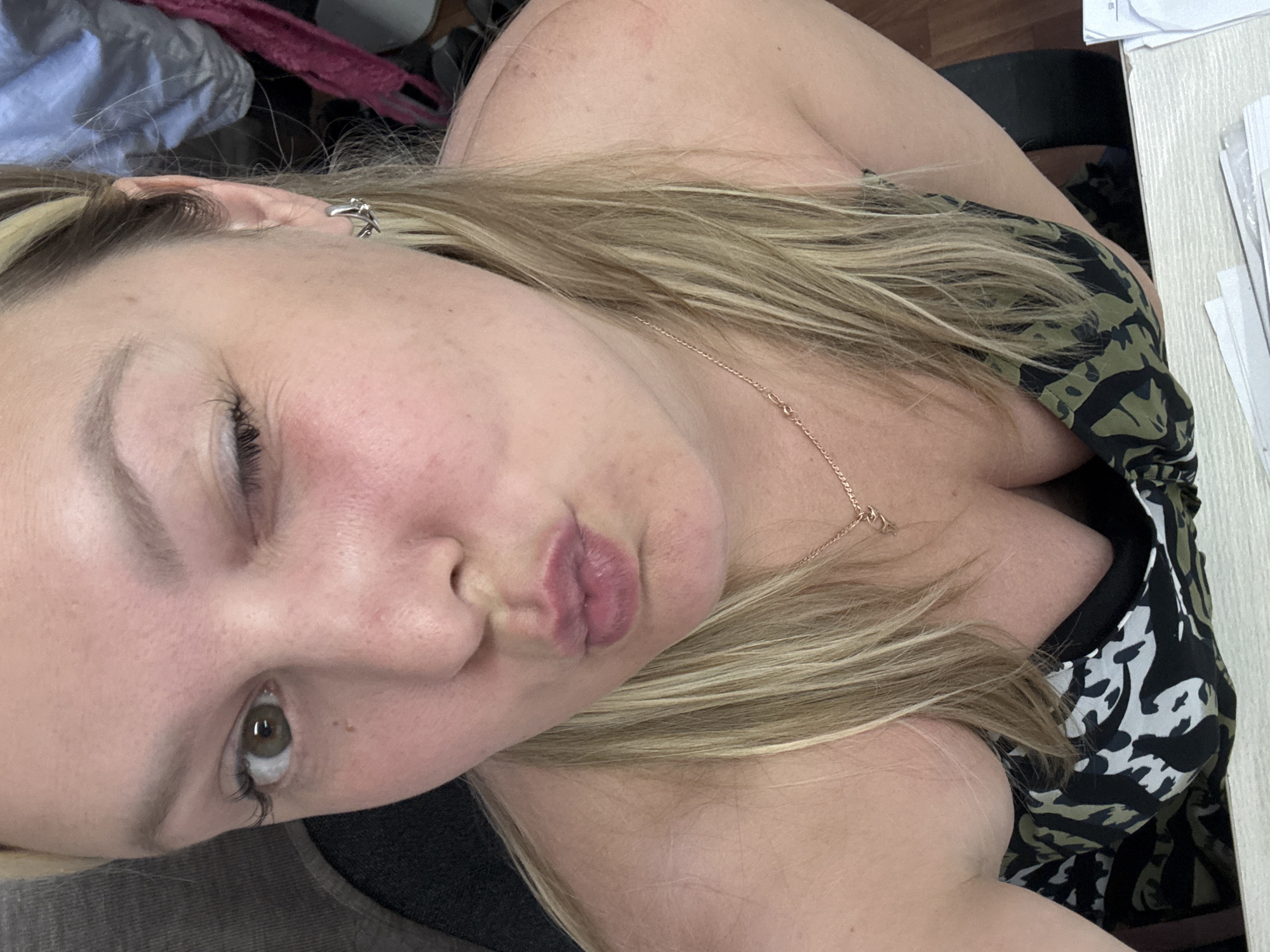 The image shows a close-up, high-angle selfie of a fair-skinned woman with blonde hair. She is wearing a black and green patterned top and a delicate gold necklace. Her left eye is open, and her right eye is closed in a wink. Her lips are pursed as if for a kiss. She is wearing a small silver earring in her visible right ear. The background is blurred but appears to be indoors, with hints of clothing and possibly furniture. The location of Noviy, Russia, is not visible in the image.