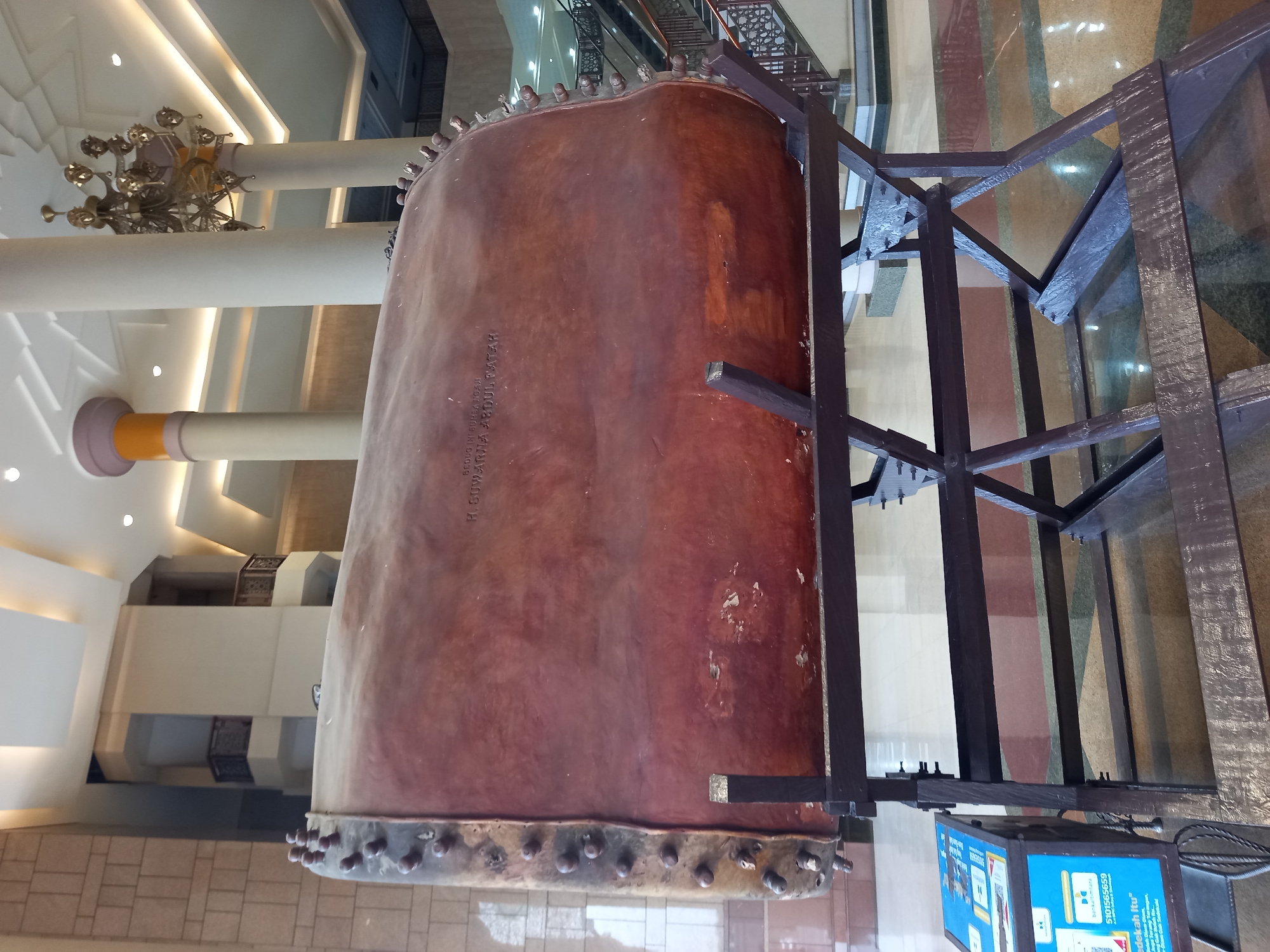 The image shows a large, wooden drum, known as a bedug, displayed on a dark wooden stand in an indoor public space. The drum is reddish-brown and appears to be made from a single piece of wood, with metal studs around its circumference. There is an inscription on the surface of the drum, which reads "BEDUG INTISURGA NOAH WARIA ABDUL FATTAH". To the left of the drum, there is a blue informational kiosk with some text and graphics. The background reveals a modern building interior with tall pillars, intricate ceiling designs, and balconies. Staircases and escalators are visible further in the background, suggesting a spacious public building such as a museum or a government complex. This bedug is located at the Masjid Islamic Center Samarinda in the city of Samarinda, East Kalimantan, Indonesia.