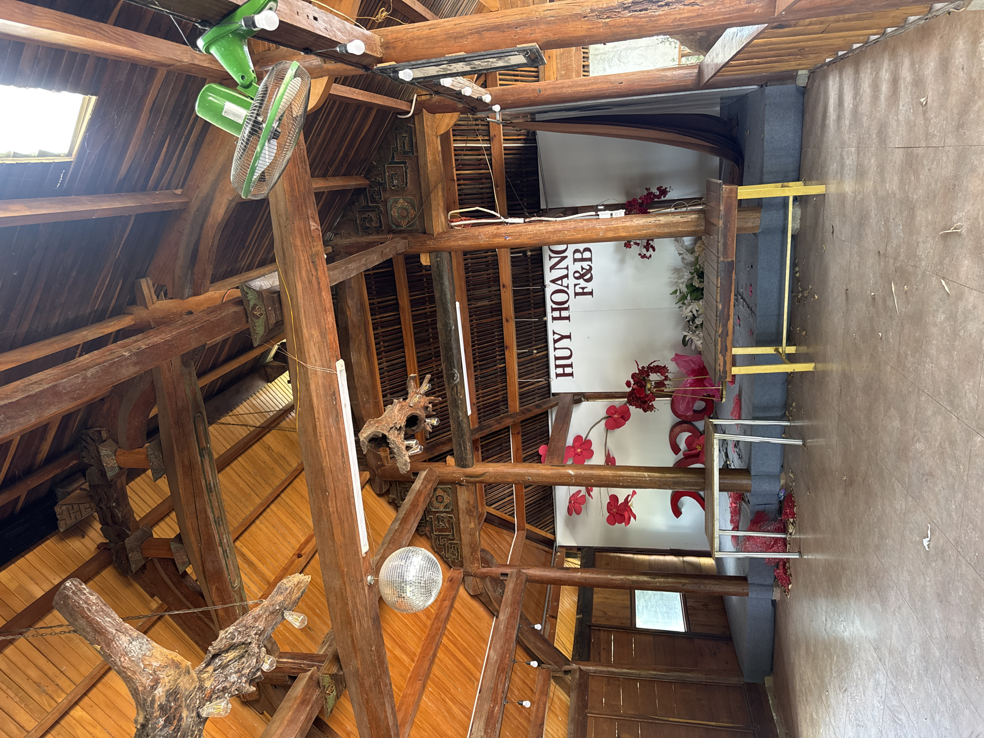 The image shows the interior of a building in Phú Thạnh, Vietnam. The ceiling is constructed of wood beams and planks, with a green fan and a disco ball hanging from it. A sign reading "HUY HOÀNG F&B" is visible against a white backdrop. Red decorations are scattered around the room. A platform or stage is also present. The floor is tiled.