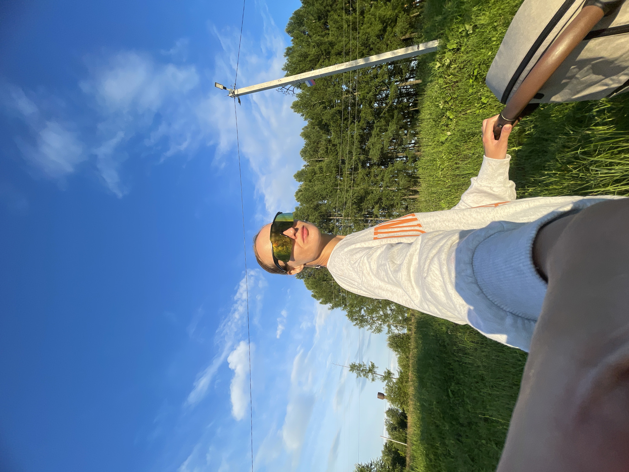 A low-angle, wide-shot photograph shows a woman from the chest down, pushing a stroller. She is wearing a light gray sweatshirt with orange lettering, large reflective sunglasses, and her hair is pulled back. She is holding the handle of a gray stroller. The background features a bright blue sky with scattered white clouds, a line of tall green trees, and a tall utility pole with a light and a small Russian flag attached to it. To the left of the utility pole, there is a water tower. The scene is set in a grassy, outdoor environment, likely a rural or suburban area. The image appears to be taken in the city of Podshivalovo, Russia, judging by the presence of the Russian flag and the description of the location. The sun is low in the sky, casting a warm light on the scene, suggesting it is either early morning or late afternoon.