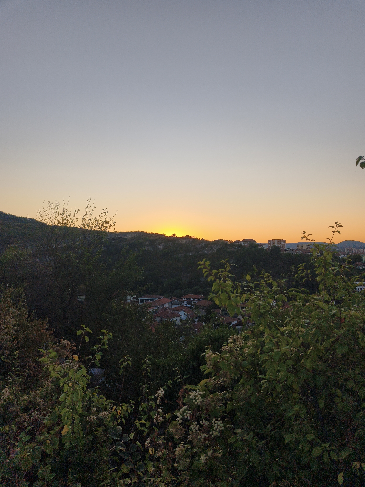 This is a landscape photograph taken at dusk, looking out over the city of Lovech, Bulgaria, from a vantage point above the town. The sky is a gradient of pale blue at the top, transitioning to a vibrant orange and yellow near the horizon, indicating the setting sun. In the mid-ground, a dense line of trees and a hillside with rocky outcrops obscure the lower portion of the town. A cluster of buildings with red tile roofs is visible nestled amongst the greenery on the slopes. Further in the distance, more buildings of varying heights and architectural styles can be seen, some with lighter colored facades. The foreground is dominated by the dark silhouettes of trees and foliage, with some branches and leaves clearly defined against the softer light of the background. The overall atmosphere is tranquil and serene, characteristic of a sunset view over a city. There are no people or animals visible in the image.