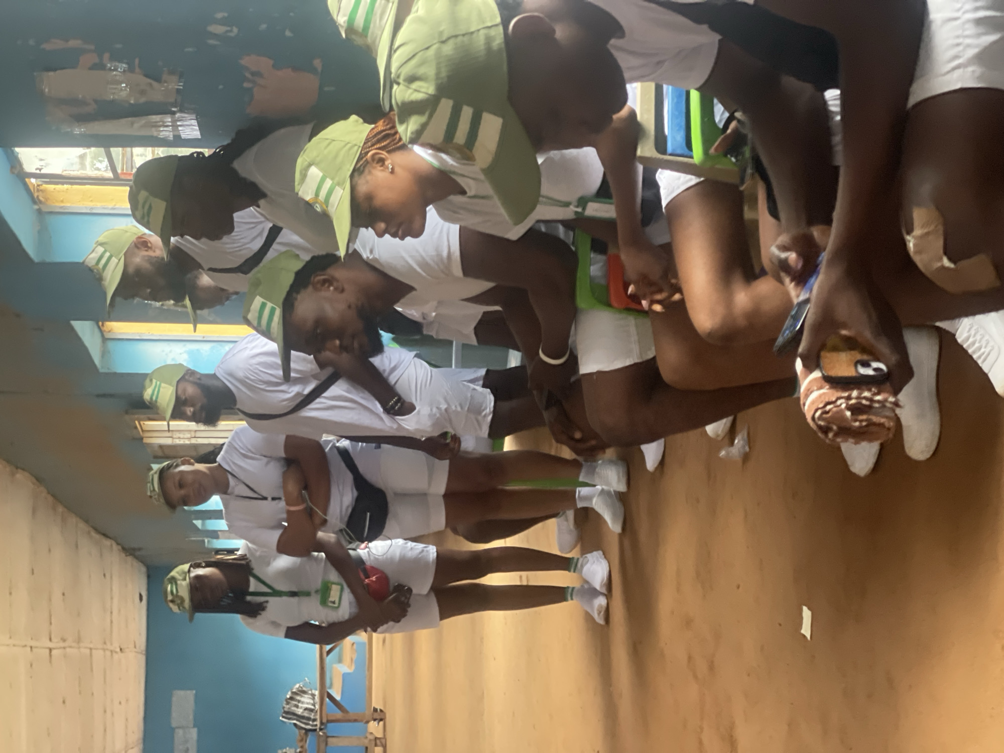 In Ise-Ekiti, Nigeria, a group of young people, likely participants in a youth service program given their matching white t-shirts, white shorts, and green caps with a white emblem, are gathered indoors. The room appears to be a classroom or hall with light blue painted walls and a textured ceiling. Natural light streams in from windows on the right side of the frame, illuminating dust particles in the air. The floor is a reddish-brown, possibly compacted earth or dirt. Several individuals are seated on colorful plastic chairs, while others stand. Some are looking at their mobile phones, suggesting a moment of downtime. One person in the foreground is holding a phone with a decorative phone case. Another individual is seen with a bandage on their knee. The overall atmosphere seems relaxed, with the young people in a communal setting.