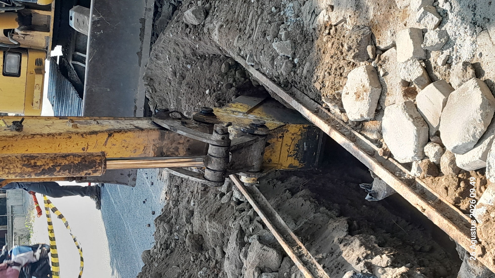 An excavator is digging a trench on a street in Takeran, Indonesia. The excavator is yellow and has a large metal bucket. The trench is deep and filled with dirt and rocks. There are yellow and black caution tapes in the background. A person wearing dark pants and boots is standing near the excavator. The image is dated August 26, 2026.