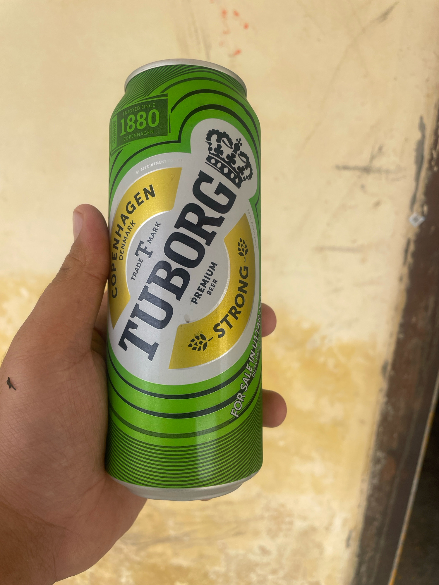 A hand holds a green and silver can of Tuborg Strong beer. The can is decorated with a crown and the brand name. The background is a light-colored, textured surface, possibly a wall or table. The location is specified as Rudrapur Elaquakaholiya, India.