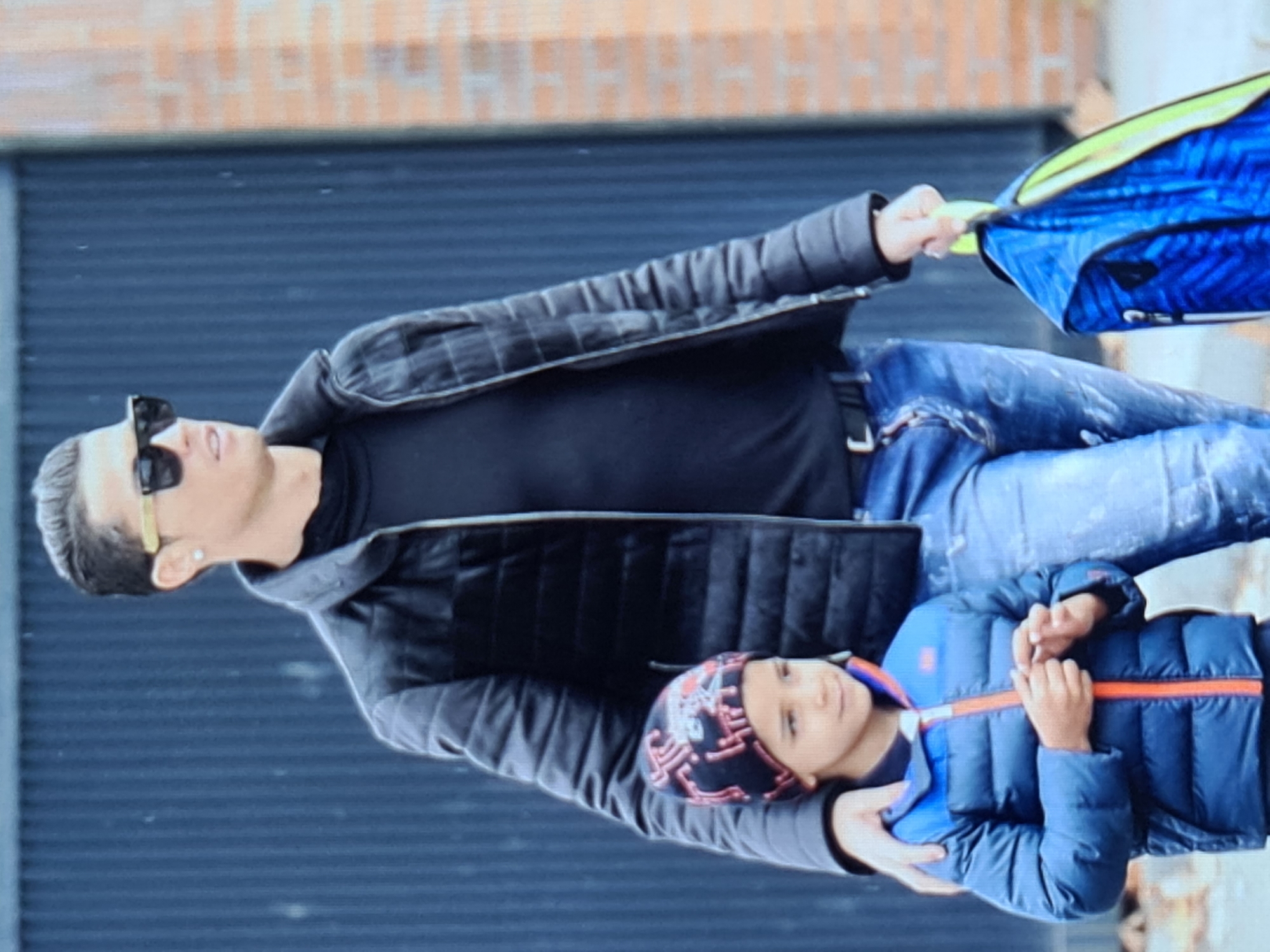 The image shows a man and a young boy walking outdoors, with a dark vertical fence and a brick wall in the background. The man, who appears to be Cristiano Ronaldo, is wearing a black puffer jacket over a black turtleneck and blue jeans. He has on gold-rimmed sunglasses and a diamond earring. The boy is wearing a blue puffer jacket over a dark beanie with a red and black design. He is holding the man's arm. The man is carrying a blue backpack with a yellow trim. The scene appears to be in an urban environment, possibly on a street in Hrazdan, Armenia. The lighting suggests it is daytime, and there might be light rain or mist, as indicated by small spots on the fence.