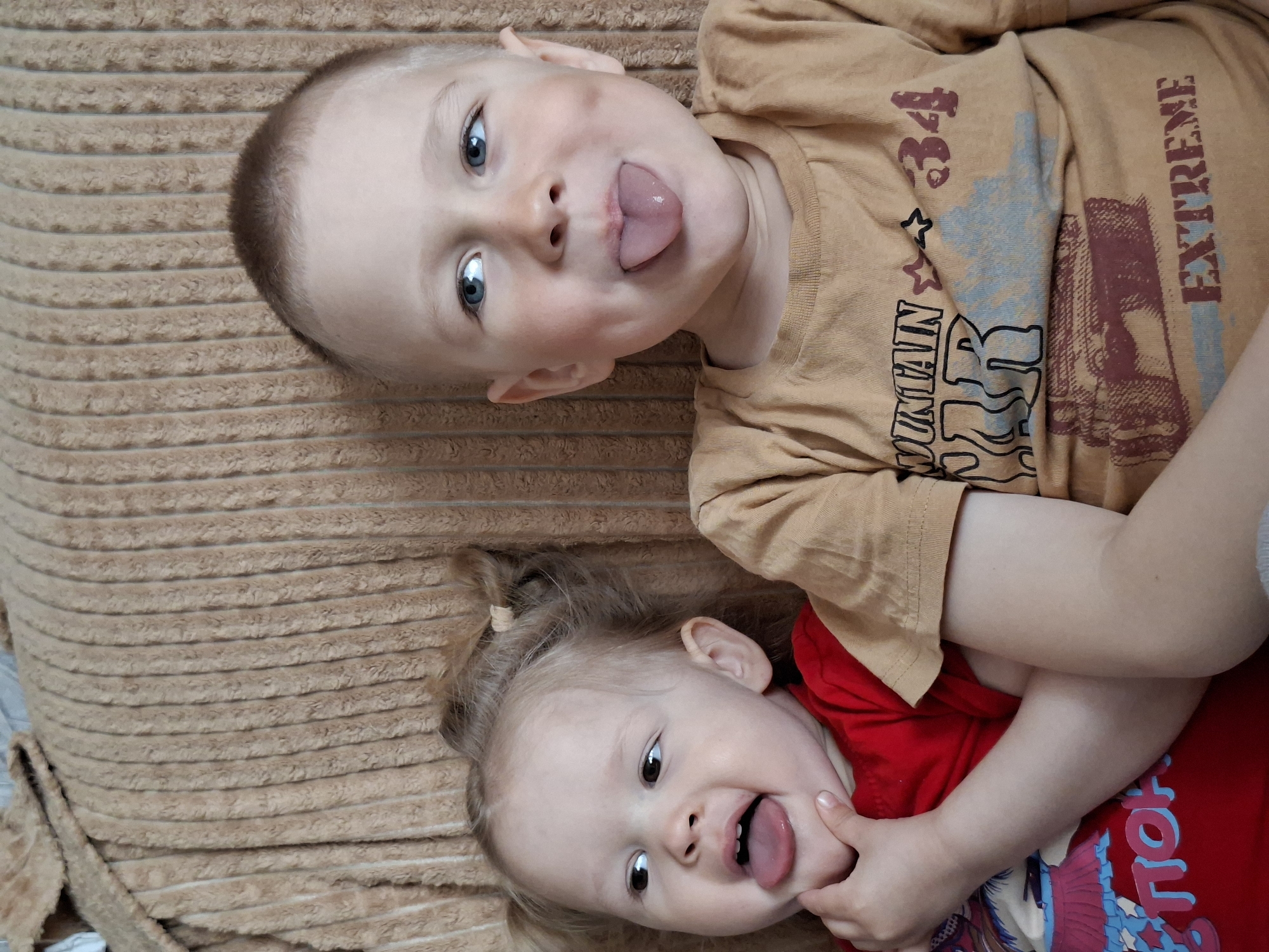 Two young children are lying on a brown, corduroy-textured surface. The child on top is wearing a tan t-shirt with the words "MOUNTAIN" and "EXTREME" printed on it, and is sticking their tongue out at the camera. The child below is wearing a red t-shirt and is also sticking their tongue out. They are holding their cheek with one hand. Their bodies are intertwined, with the top child's arms wrapped around the bottom child. The location appears to be indoors, possibly on a bed or sofa.