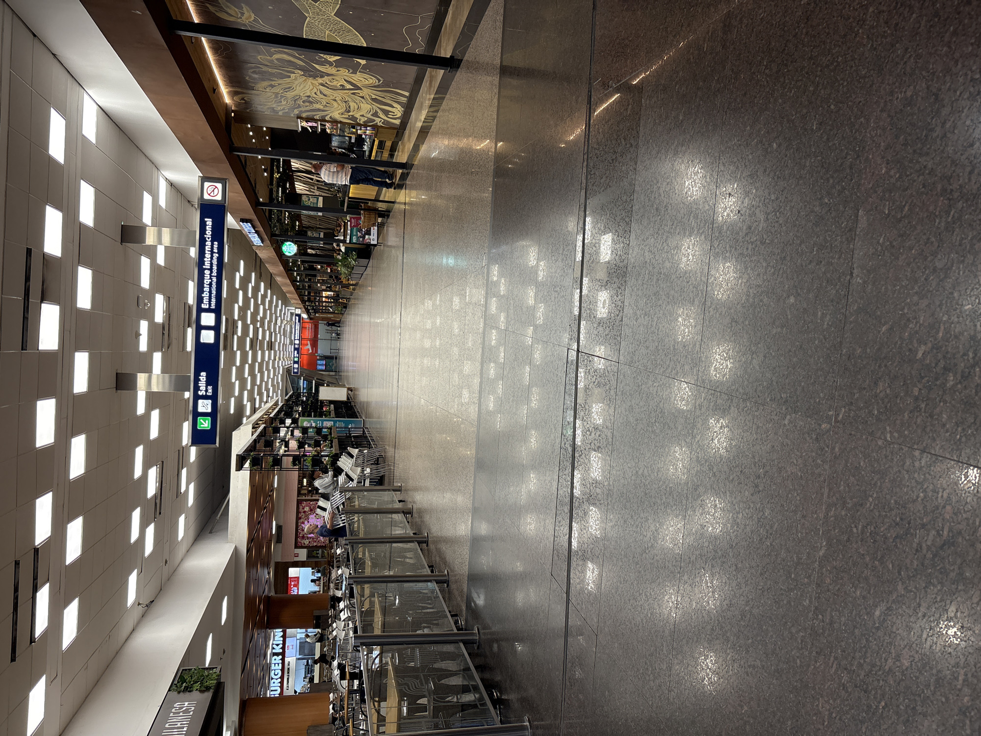 The image displays an interior view of a wide, brightly lit concourse within Ministro Pistarini International Airport (EZE) in Buenos Aires, Argentina. The perspective is longitudinal, looking down the length of the hall.
The overhead ceiling is white, featuring a grid of evenly spaced square recessed fluorescent light fixtures that cast strong reflections onto the highly polished, dark grey or dark brown tiled floor. Black strips delineate sections of the floor.
To the left, a commercial food court area is visible. A prominent "BURGER KING" sign and a partial "MILANESA" sign identify establishments. This area contains numerous light-colored tables and chairs, partially enclosed by a low barrier constructed of glass panels framed by silver-colored metal posts. Several individuals, approximately 5-7, are observed seated or standing within this dining section.
To the right, another commercial strip includes a "STARBUCKS COFFEE" outlet. Adjacent to the Starbucks, a distinctive brown wall is adorned with a large, stylized gold-colored graphic, depicting what appears to be a mythological creature. One male in a striped shirt and blue jeans stands at the Starbucks counter, facing away from the viewer.
Overhead, blue rectangular signs with white text and symbols indicate "Salida" (Exit) and "Embarque internacional" (International boarding area). A circular "No Smoking" symbol is also present. Green vertical plant installations are integrated into the decor on both sides of the concourse. In the distance, the concourse extends with additional commercial storefronts and several indistinct figures walking.